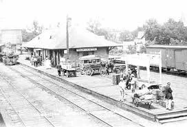 A black and white photo of a train station platform with people, a horse-drawn carriage, and train cars in the background.