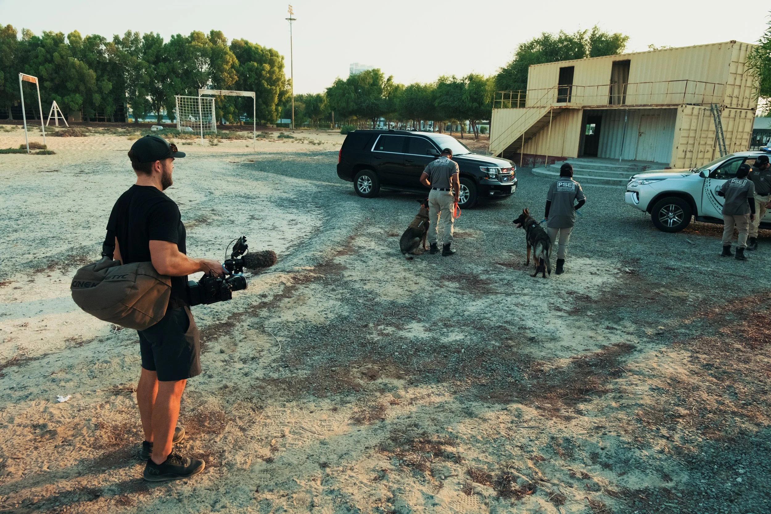 Tom Thessman - Group of law enforcement officers with police dogs in an outdoor area with parked cars, a container building, and sports fields in the background.