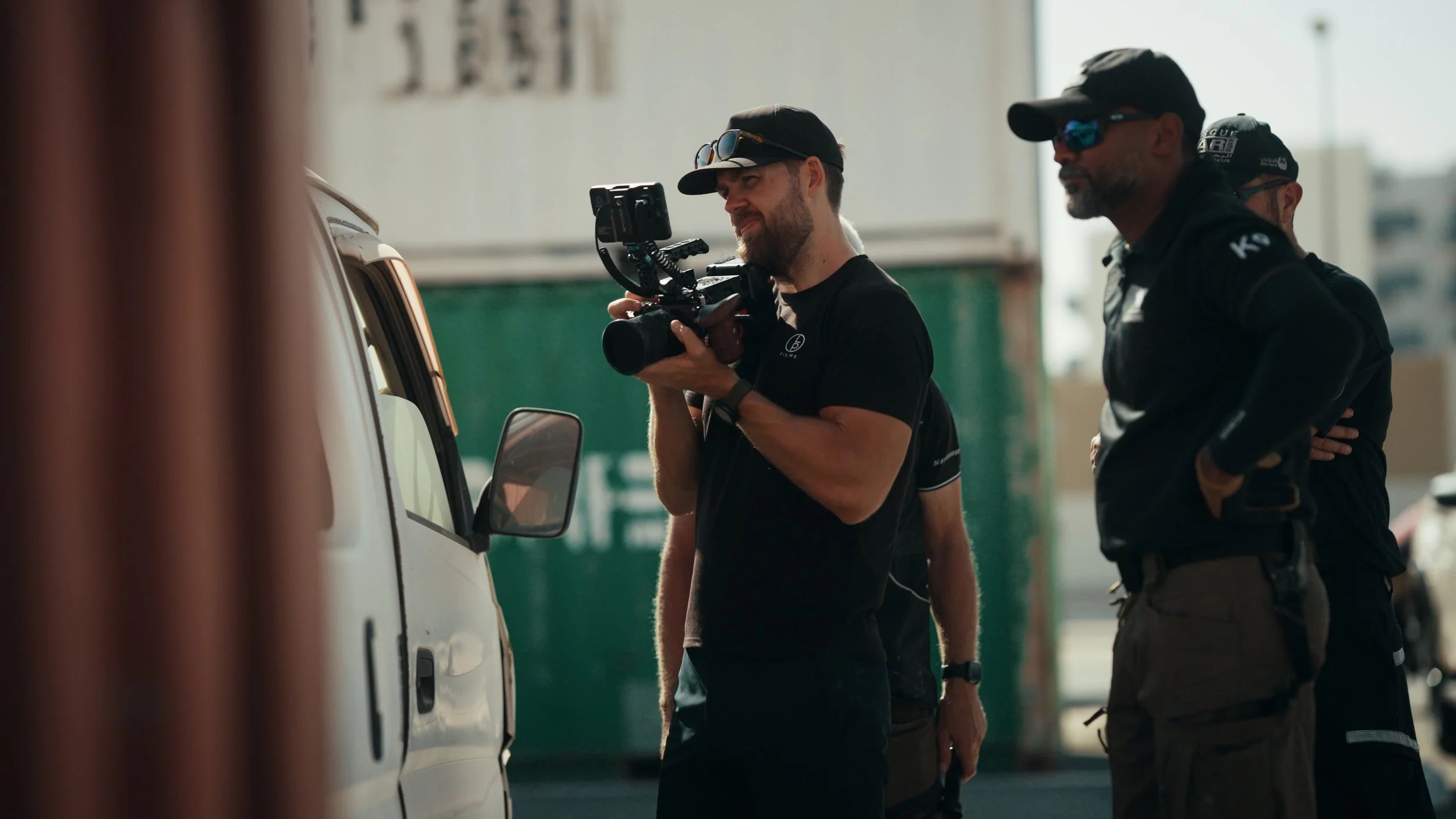 A man with a beard and sunglasses holding a camera, standing next to a white van, with two other men in dark clothing and hats nearby.