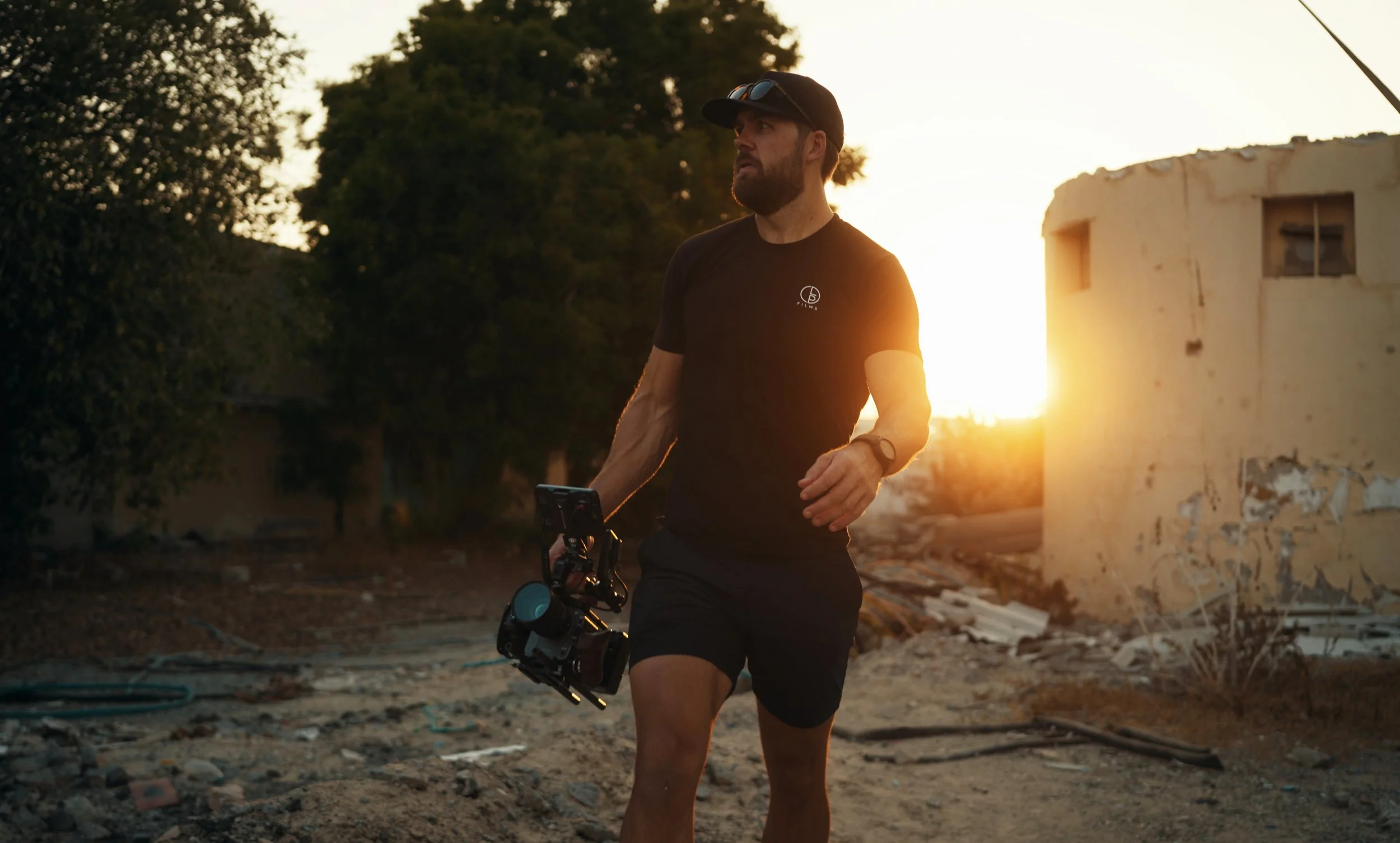 A man with a beard wearing a black cap, sunglasses on his head, and a black T-shirt running or walking outdoors during sunset, with old, damaged buildings and trees in the background.