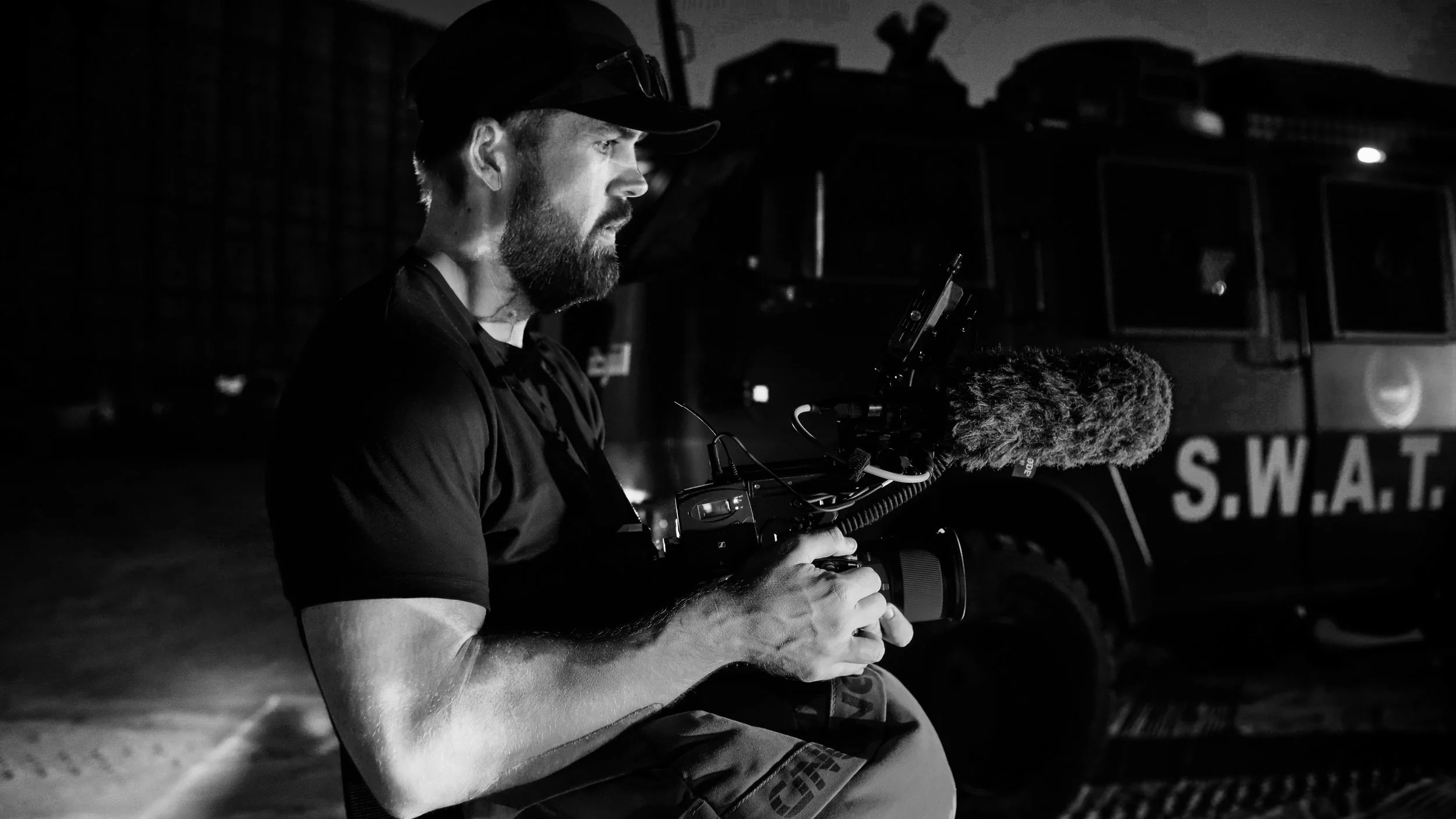 Tom Thessman - A man with a beard and wearing a cap operates a camera with a shotgun microphone attached, sitting outdoors at night with a SWAT vehicle in the background.
