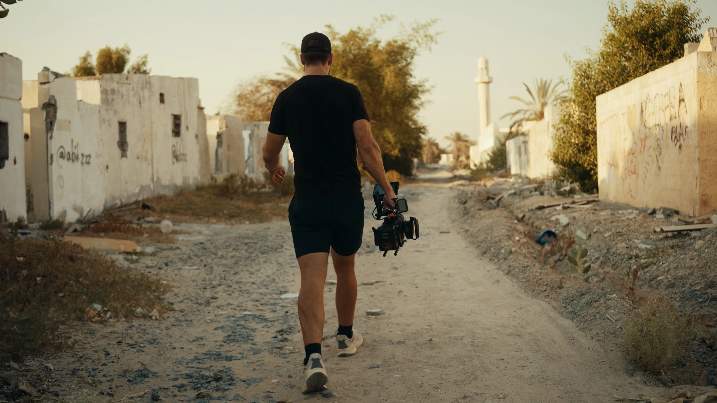 A person walking down a dirt road in a war-torn neighborhood, carrying filming equipment in one hand, with damaged buildings and graffiti on the walls.