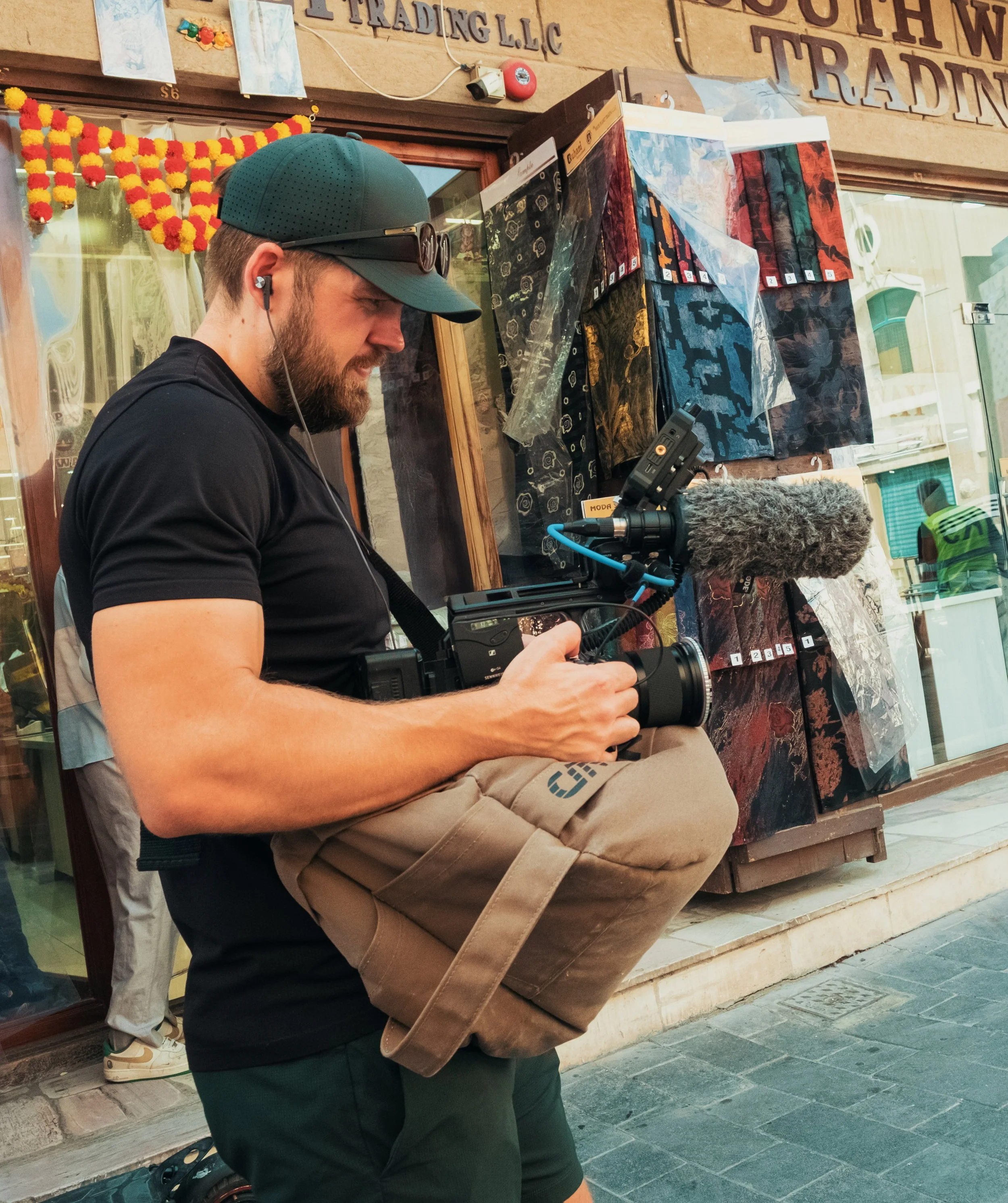 Tom Thessman - A man with a beard wearing a dark baseball cap, black t-shirt, and headphones holding a camera with a microphone on a street outside a storefront.
