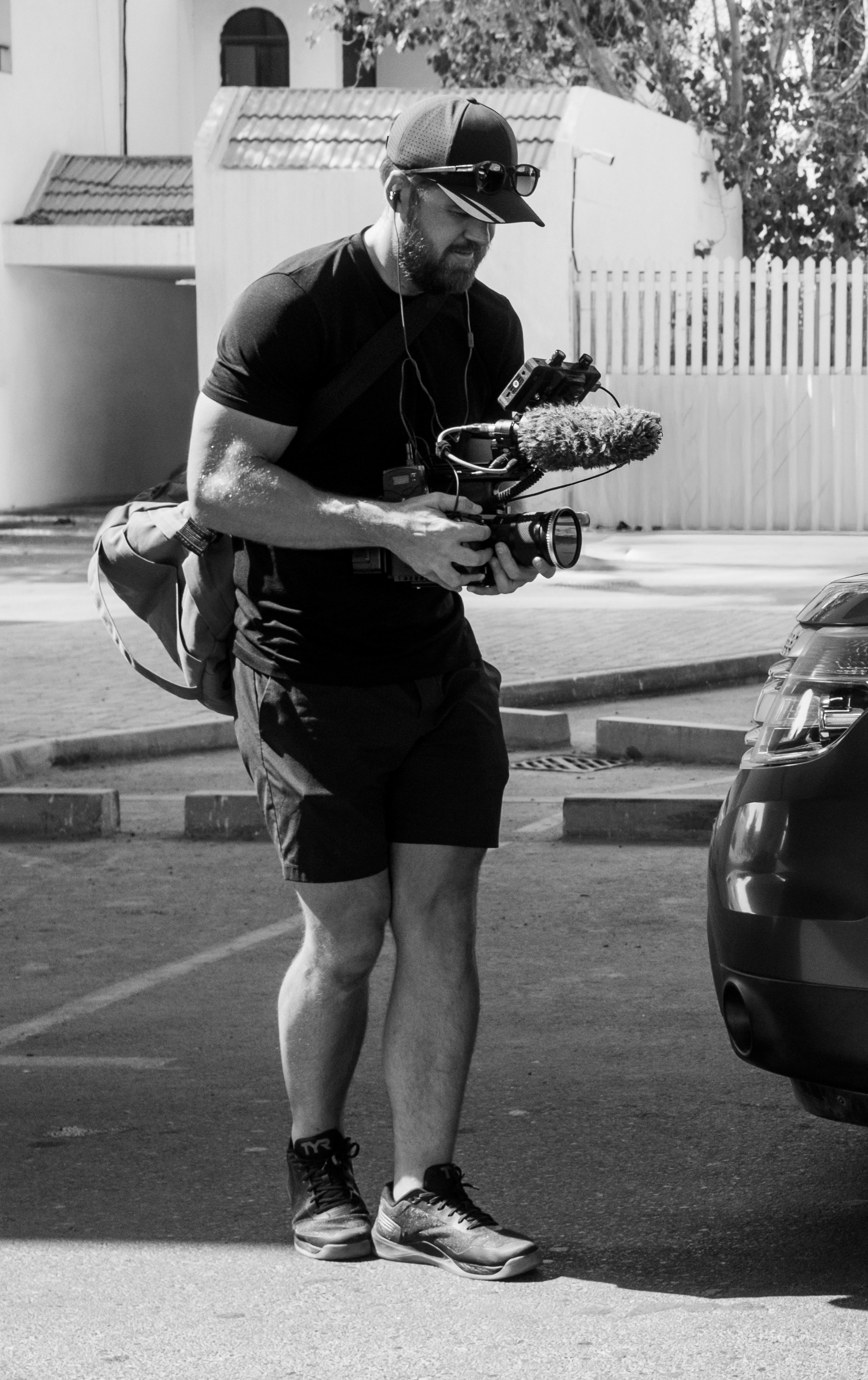Tom Thessman - A man operating a professional camera with a microphone attached, standing outdoors near a car, wearing a cap, sunglasses, t-shirt, shorts, and sneakers.