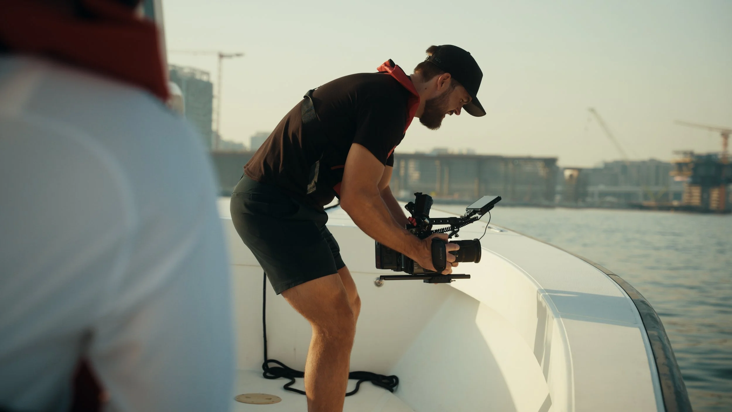 A man with a beard and wearing a black cap, black t-shirt, and shorts, operating a camera mounted on a stabilizer on a boat, with a cityscape and construction cranes in the background.