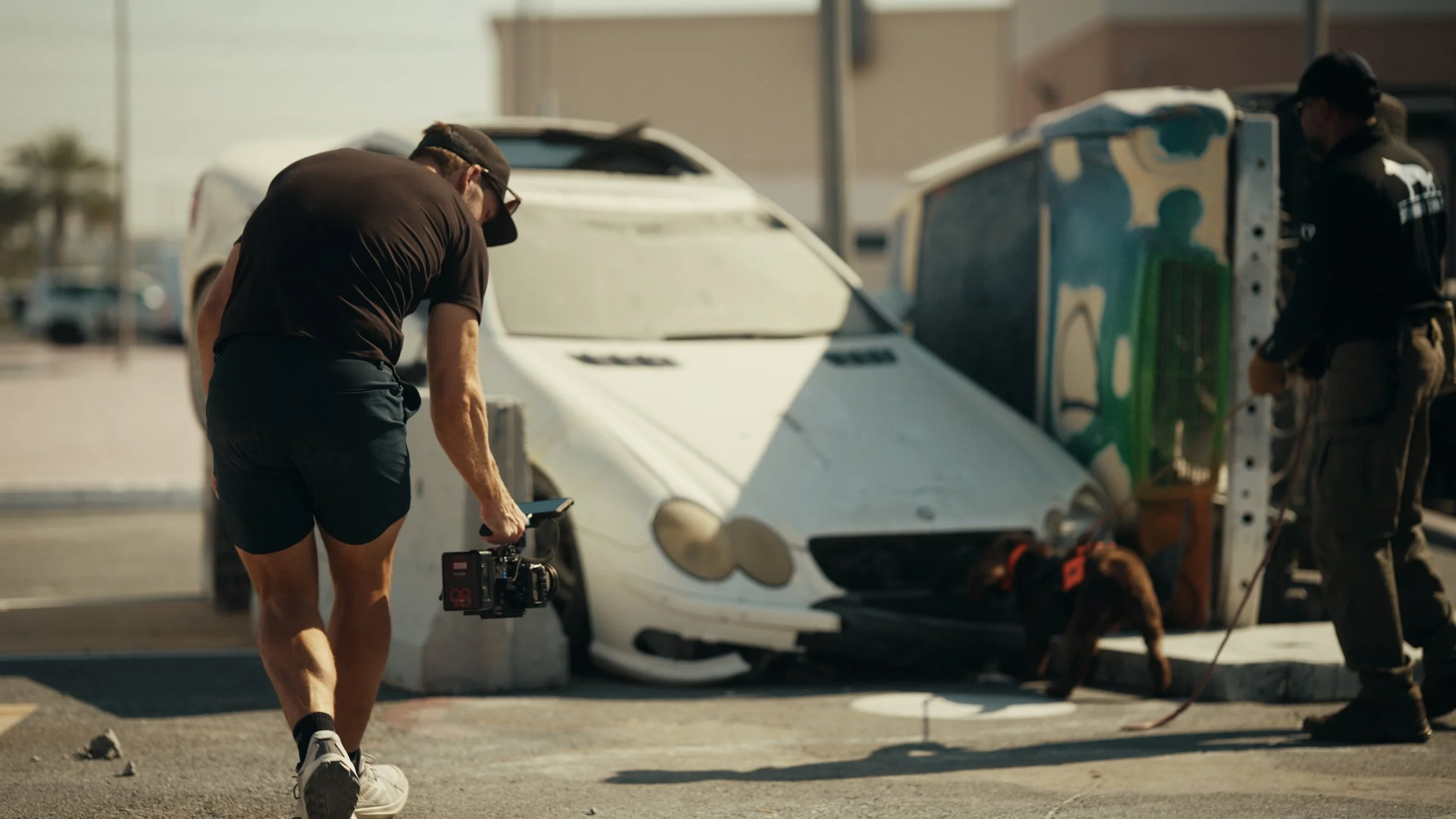 A person filming a scene with a camera in front of a vintage race car, while another person with a dog stands near a piece of equipment, with a parking lot and buildings in the background.