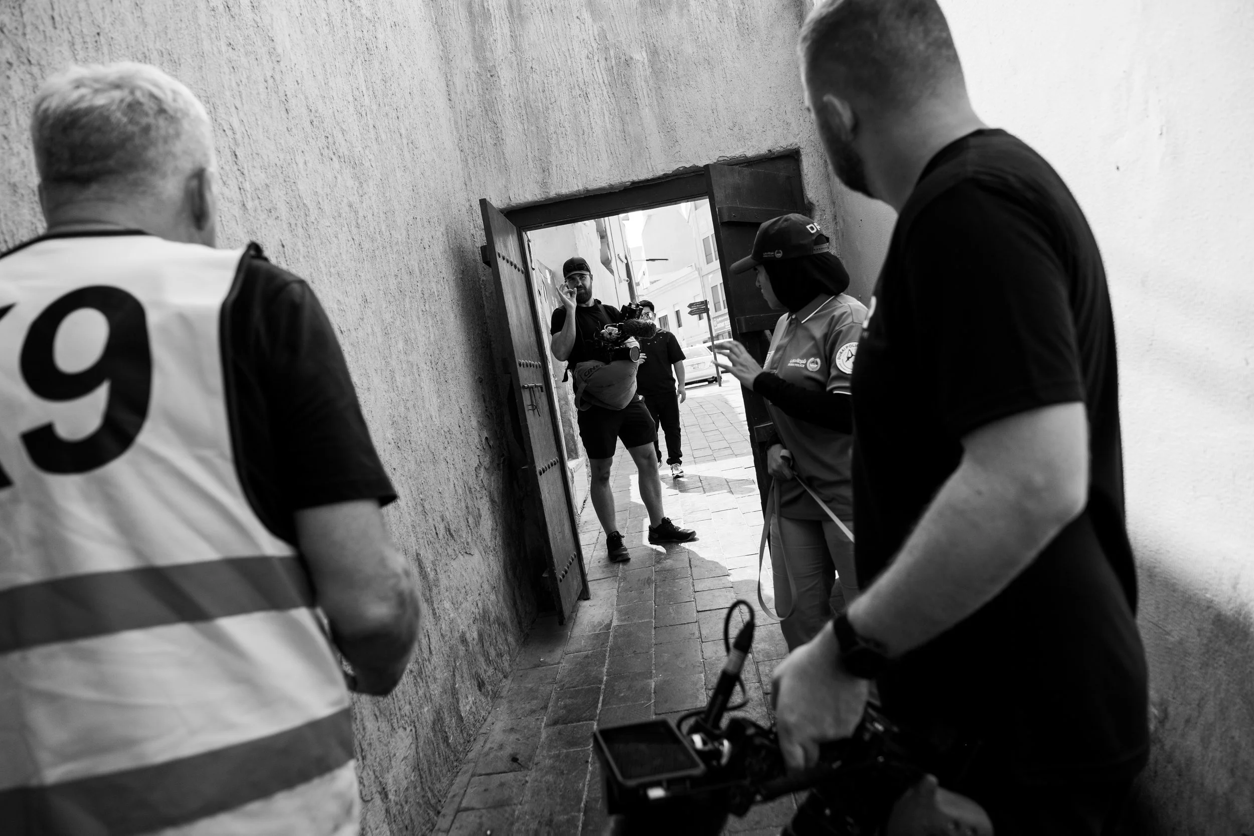 Tom Thessman - Four people standing in a narrow alleyway, with one person walking out of a doorway talking on a cellphone, and the others observing or taking notes.