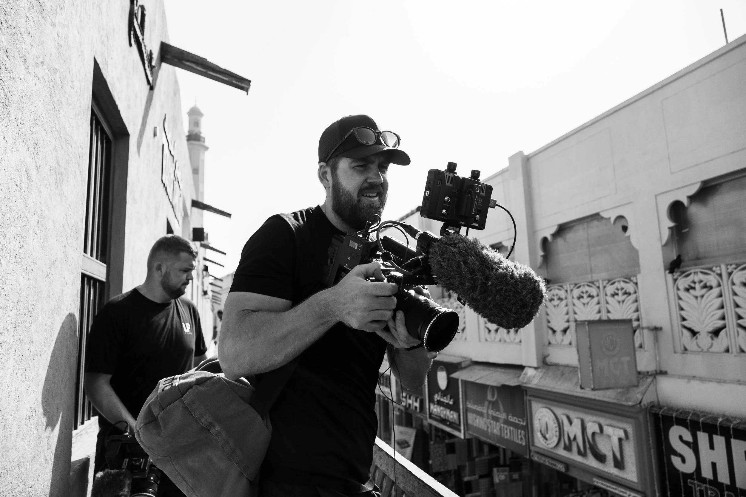 Tom Thessman - A man operating a professional video camera with a microphone in an outdoor setting, with another man standing nearby and various commercial signs on buildings in the background.