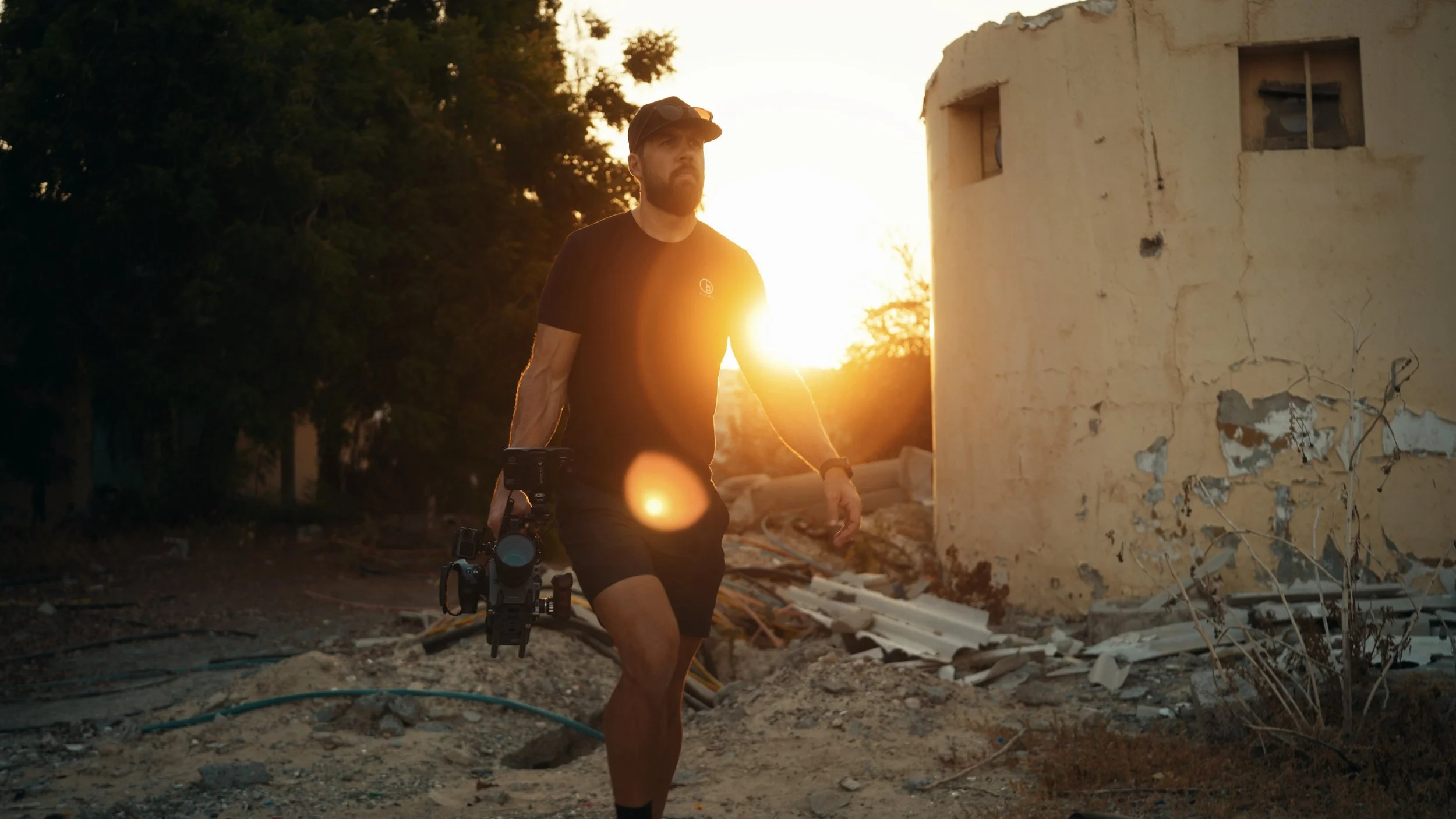 A man with a beard, wearing a black t-shirt, shorts, and a cap, walking amidst debris and damaged buildings during sunset, holding a camera.