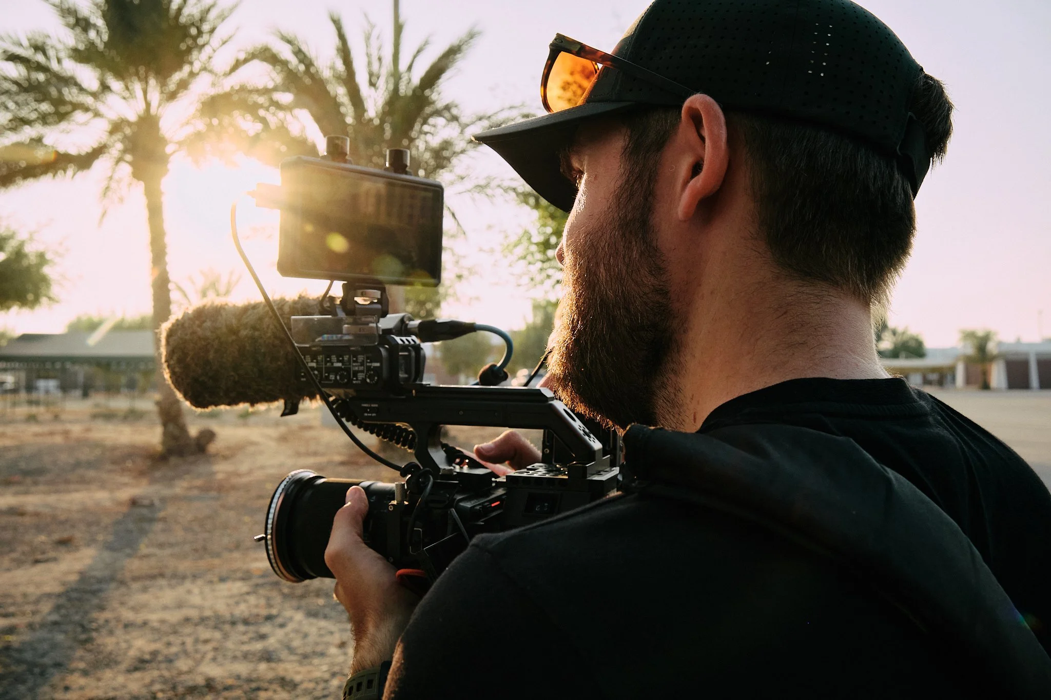 Tom Thessman - A man with a beard, wearing a black cap and sunglasses, is operating a professional video camera outdoors at sunset, with palm trees in the background.