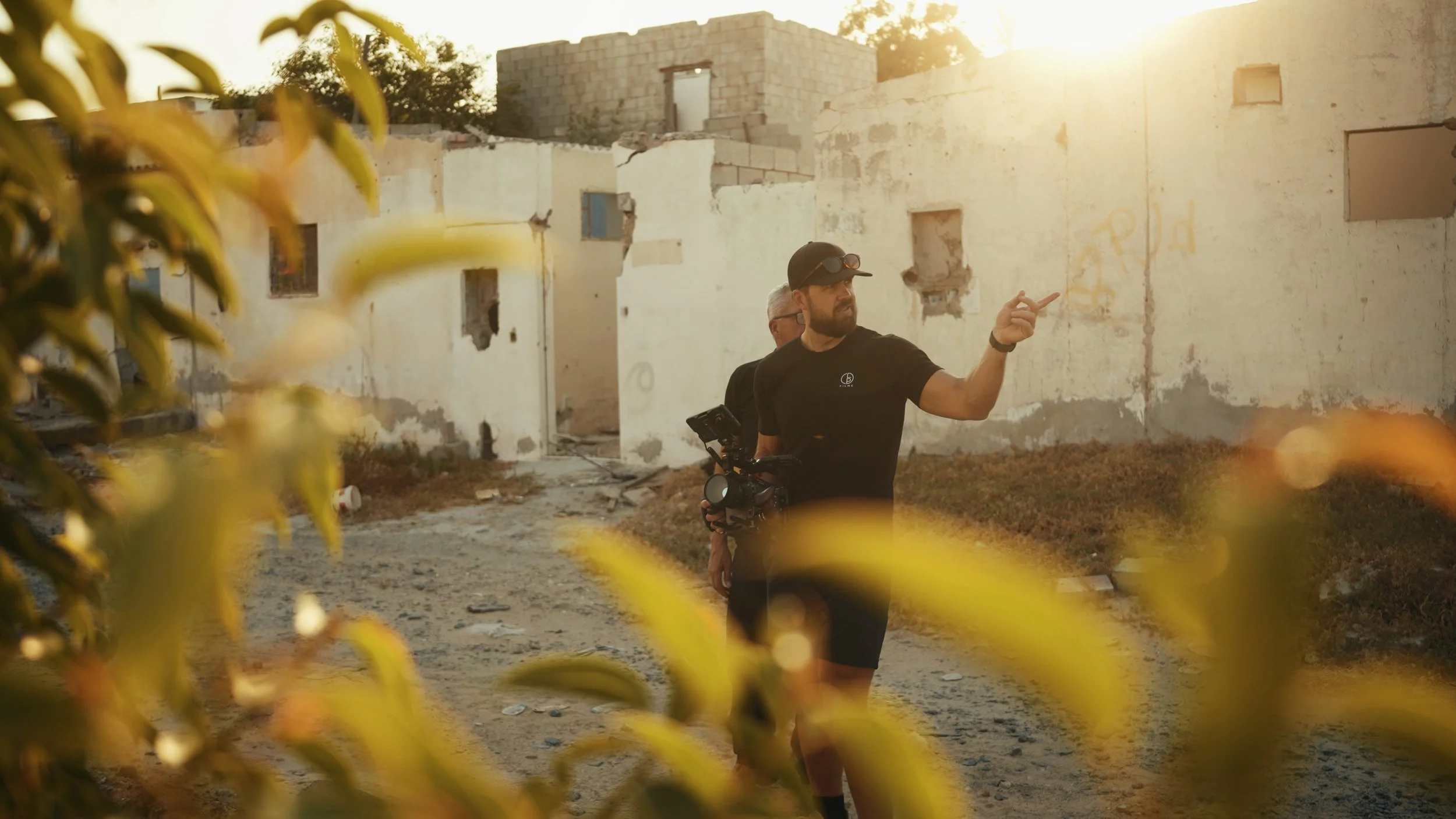 A man with a beard and cap holding filming equipment and pointing, standing outdoors in front of dilapidated white buildings during sunset, with blurred yellow leaves in the foreground.