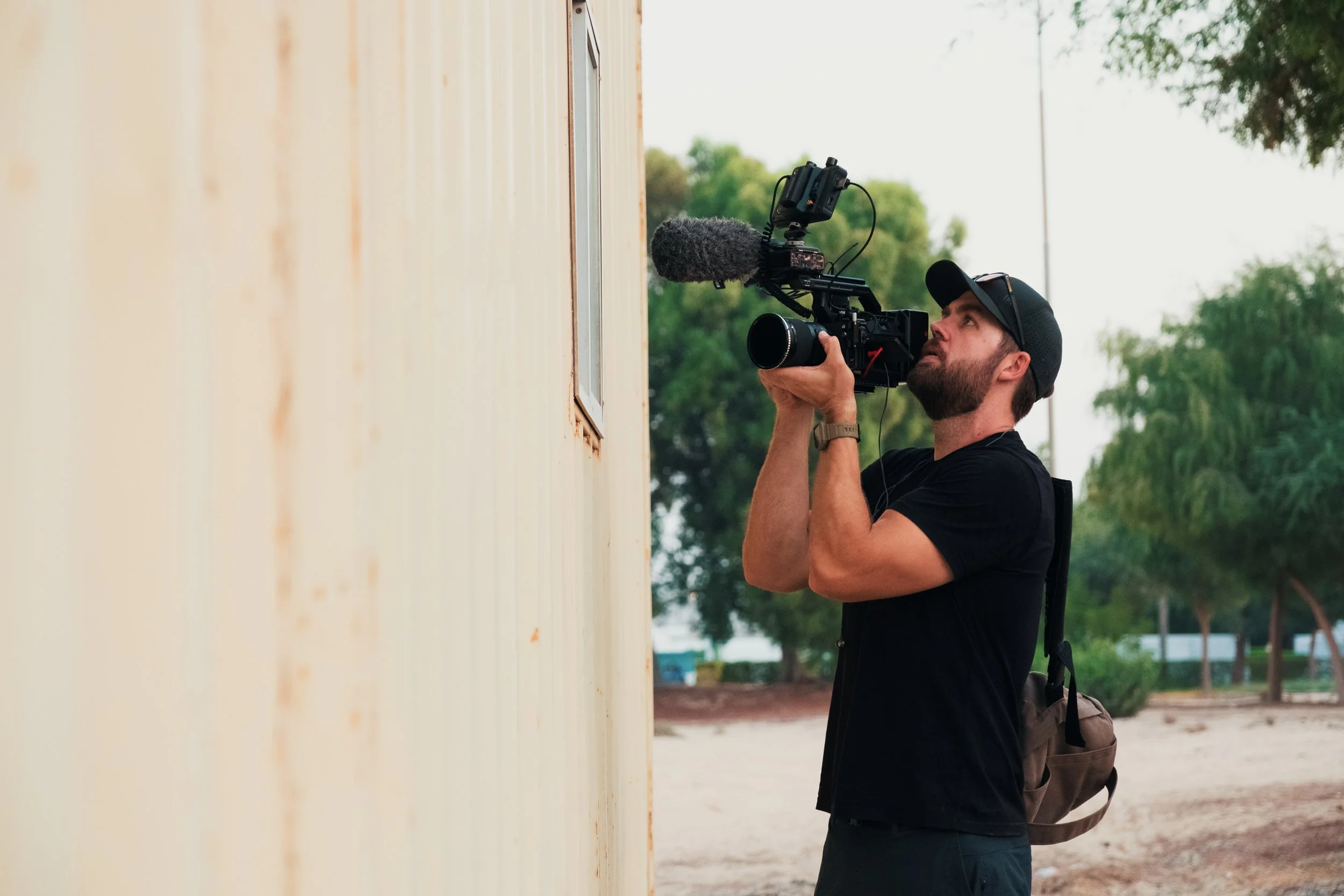 Tom Thessman - A man filming with a professional video camera outdoors next to a beige wall, with trees and a cloudy sky in the background.
