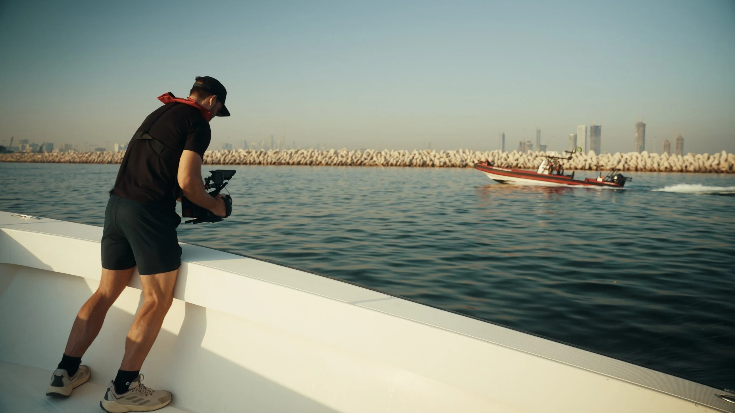 A person wearing a black cap, black shirt, and shorts is on a boat holding a camera, filming a red and black boat speeding through the water with a city skyline and breakwater in the background.