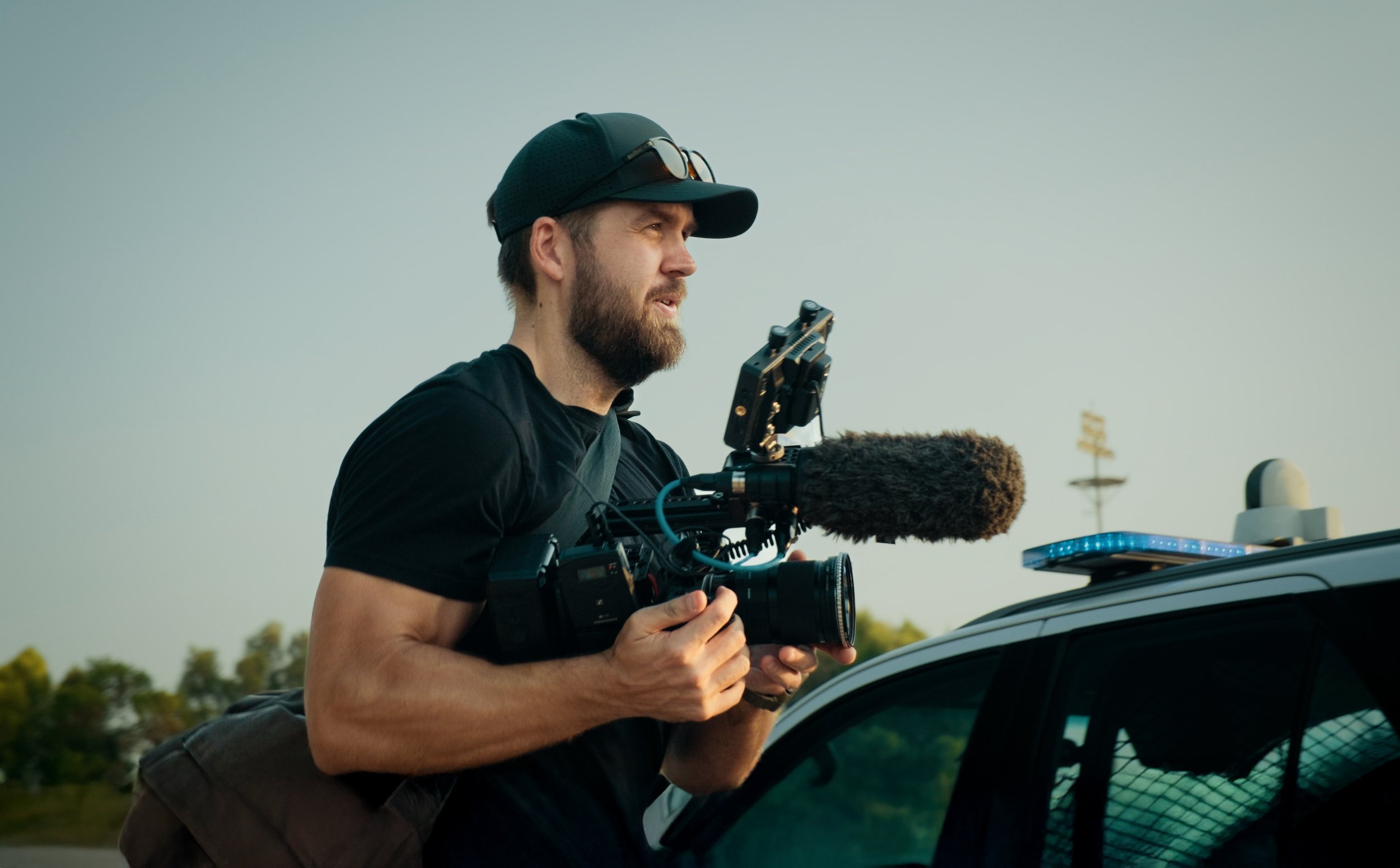 Tom Thessman - A man with a beard wearing a black cap and sunglasses on it is holding a professional camera with a microphone attachment while standing next to a police vehicle with a light bar on top during sunset or sunrise.