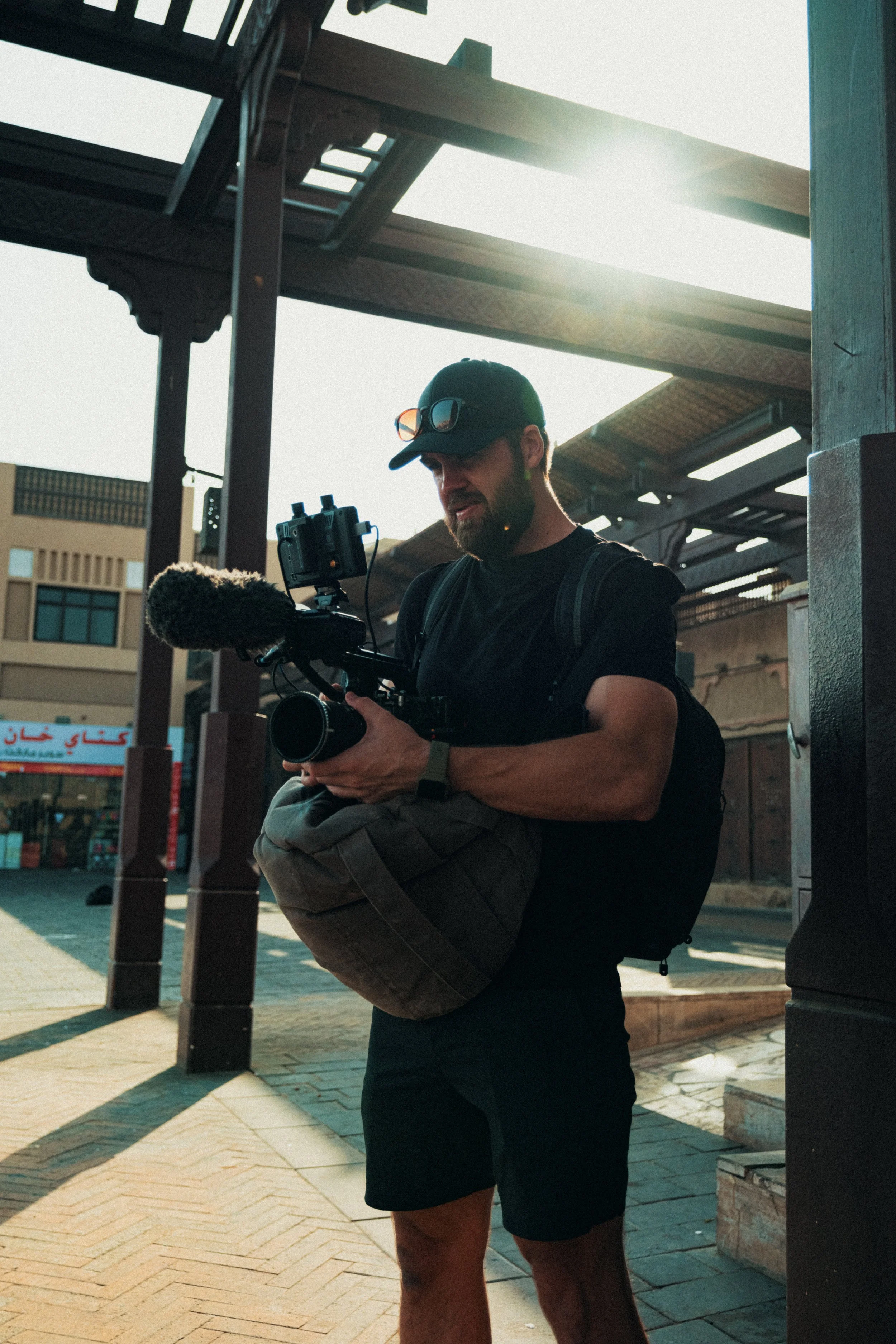 Tom Thessman - A man wearing a black cap, sunglasses, and black shirt standing outdoors during daylight, holding a professional video camera with a microphone, with a backpack, in an urban area.