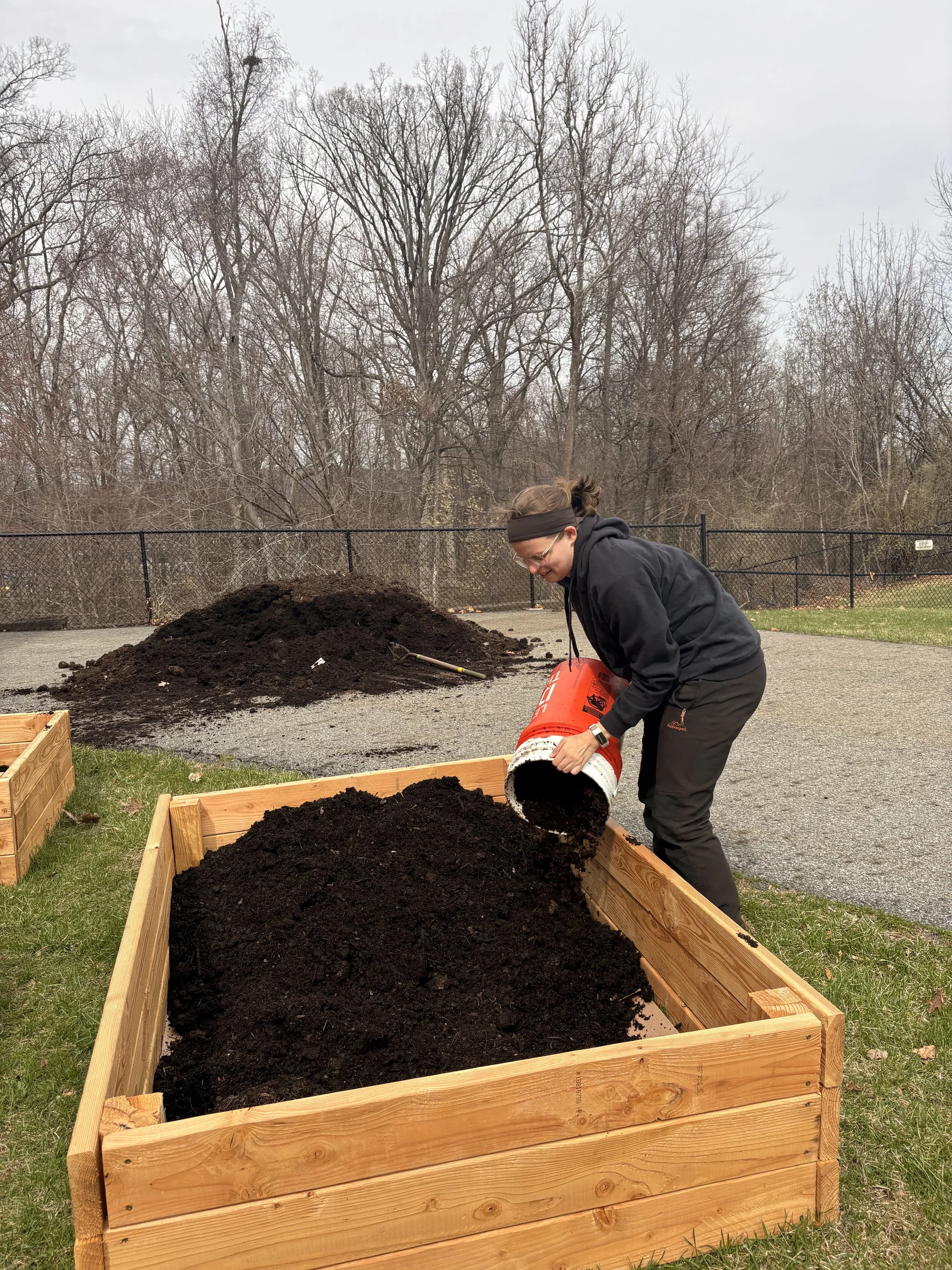 A woman in a black hoodie and sweatpants is pouring soil into a wooden garden bed. There are piles of soil and gardening tools in the background, and leafless trees can be seen in the distance.