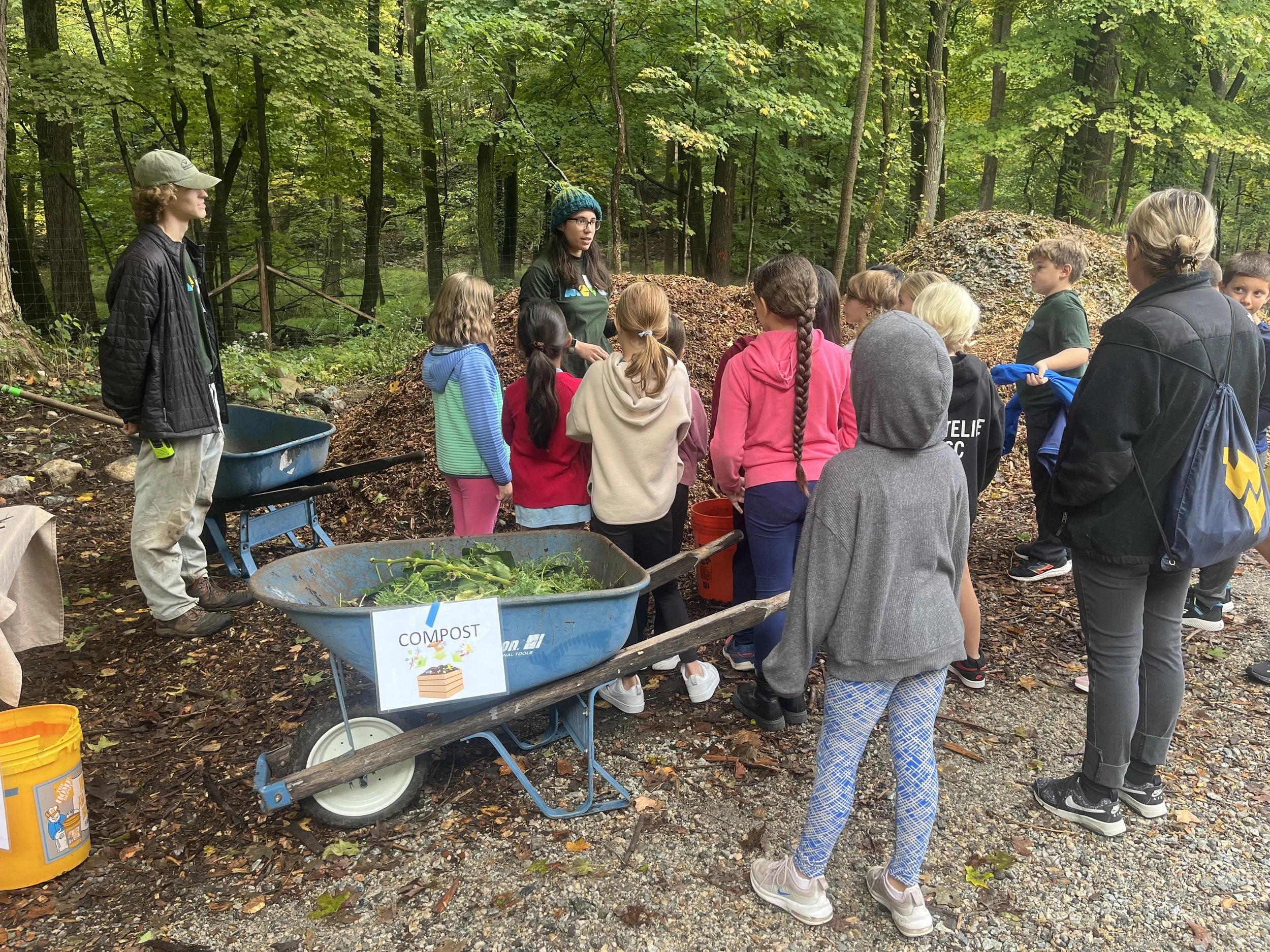 Children and adults gathered outdoors in a wooded area for an educational session about composting, with a wheelbarrow labeled "COMPOST" filled with plant materials and a sign indicating composting.