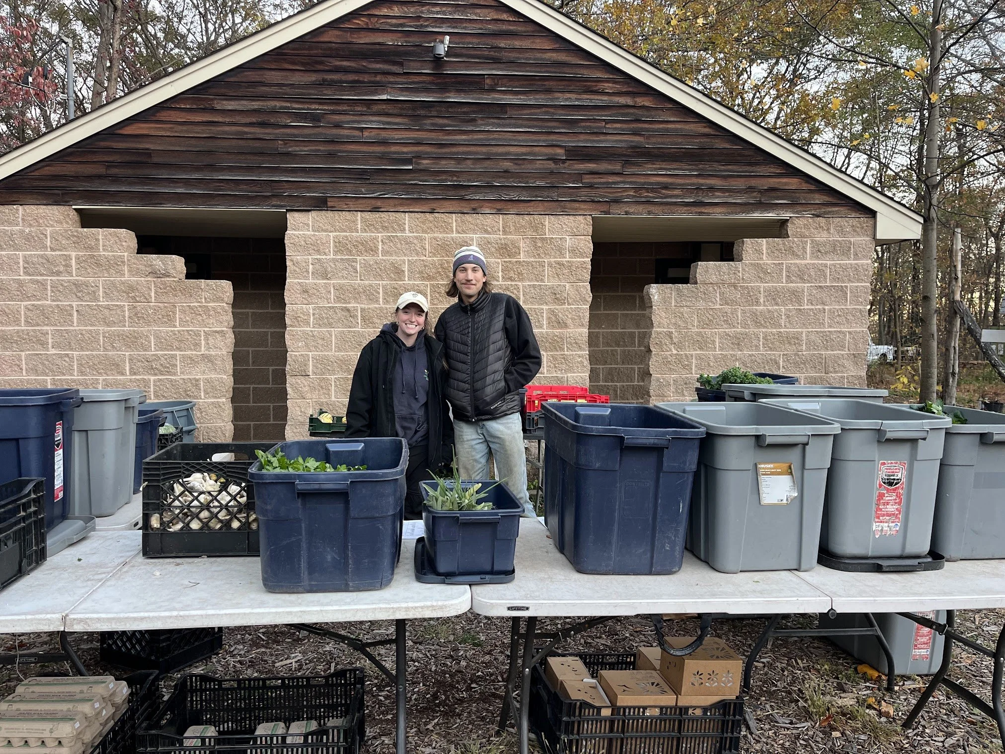 Two people standing behind a table with various bins and crates, with a brick and wood building in the background.