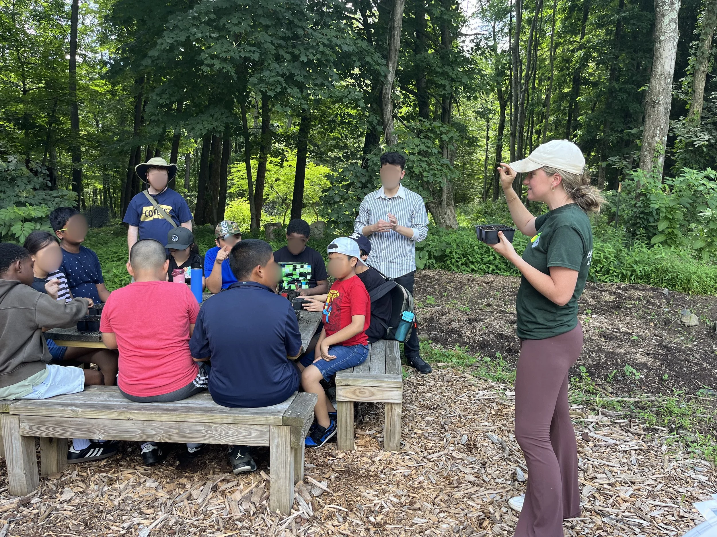 A group of children and teenagers sitting at a wooden picnic table in a forest, listening to a woman who is teaching them about plants or nature, holding a container and gesturing with her hand.