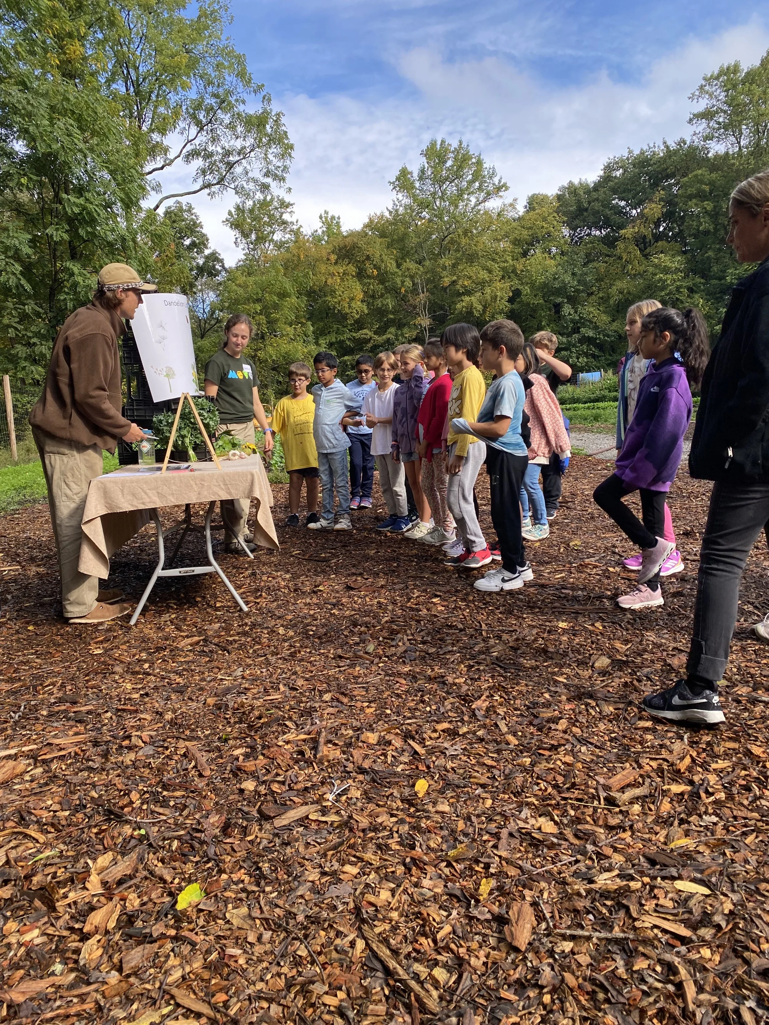 A group of children participating in an outdoor educational activity, standing on a leaf-covered ground, listening to an instructor next to a table with educational materials and visual aids, with lush trees and a partly cloudy sky in the background.