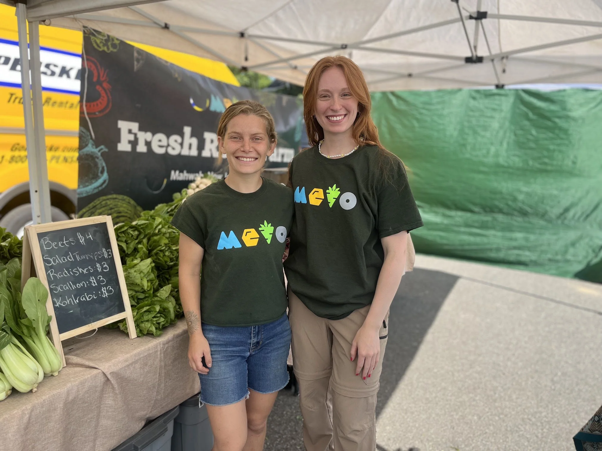 Two women standing at a farmers market stall, smiling, wearing matching mindfulness t-shirts, with fresh vegetables and a chalkboard sign listing produce prices in front of them.