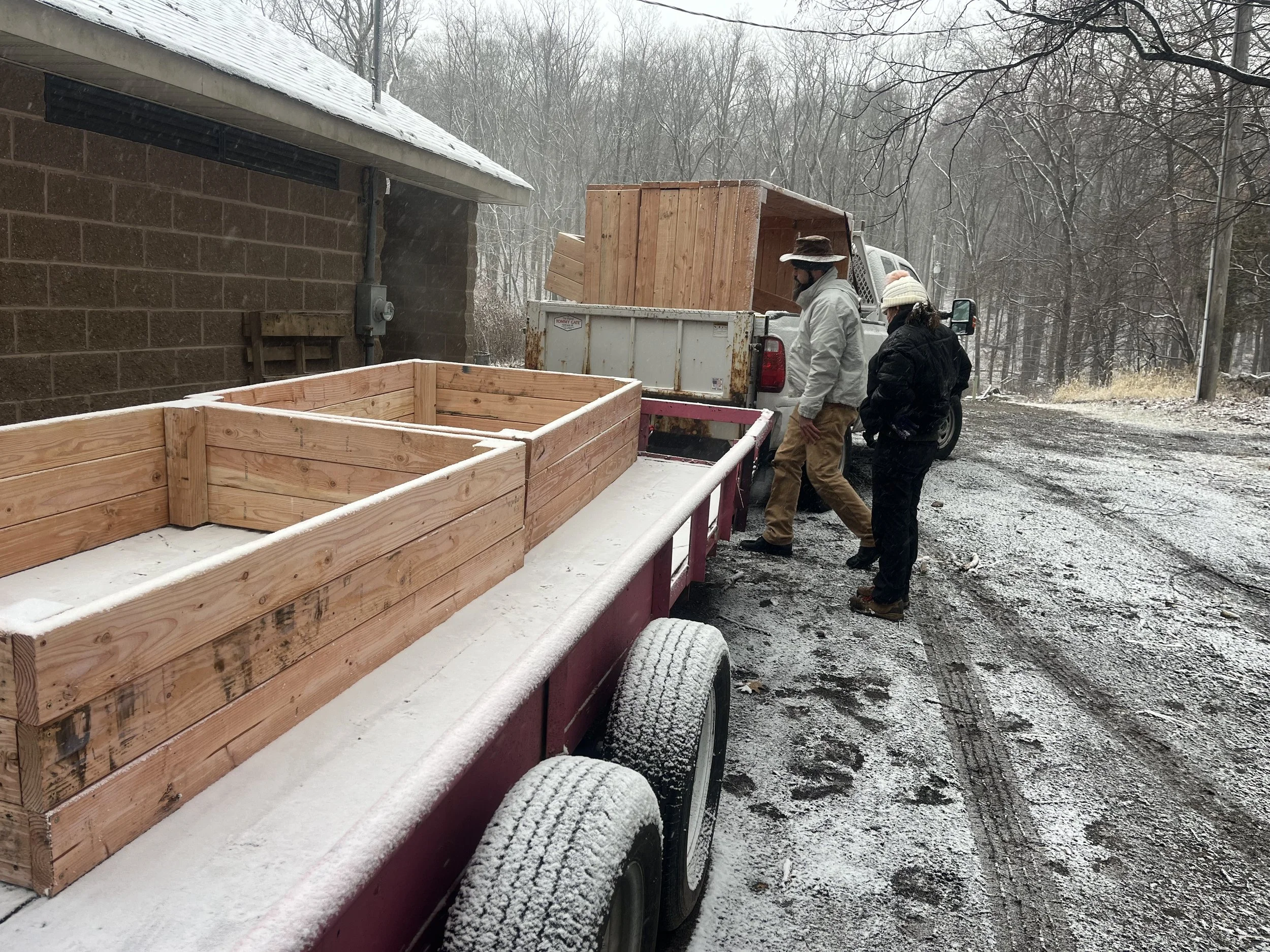 Two people loading wooden planters onto a trailer next to a building in a snowy, wooded area.