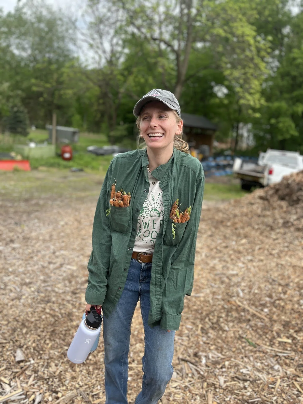 A woman in a green jacket and denim jeans stands outdoors on a gravel path, smiling and holding a water bottle. She has baby carrots tucked into her jacket pockets. She is wearing a gray cap and a t-shirt that says "Sweet Root Farm." The background features green trees, a small building, and a white truck.