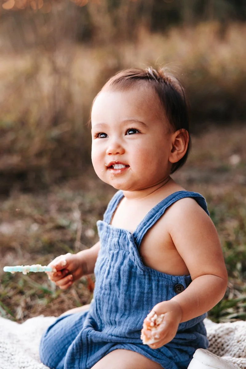 Smiling baby boy in blue suspenders eating 1st birthday cake