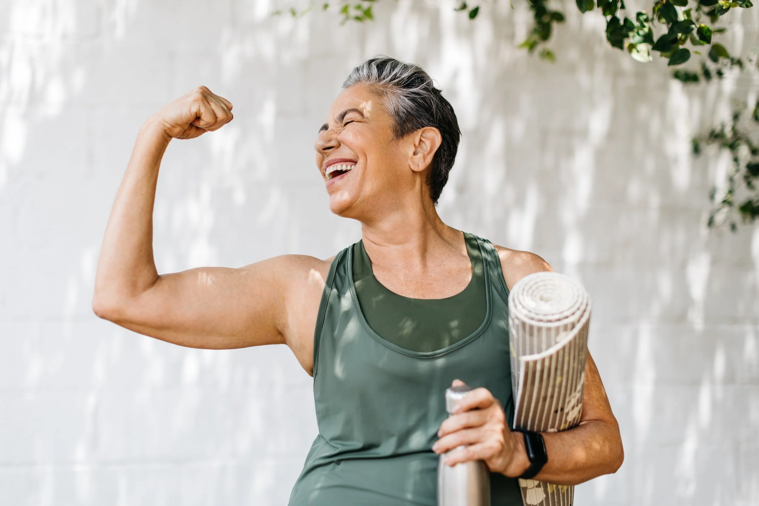 Elderly woman outdoors celebrating her fitness achievement by flexing her bicep, symbolizing strength, vitality, and longevity through midlife training.