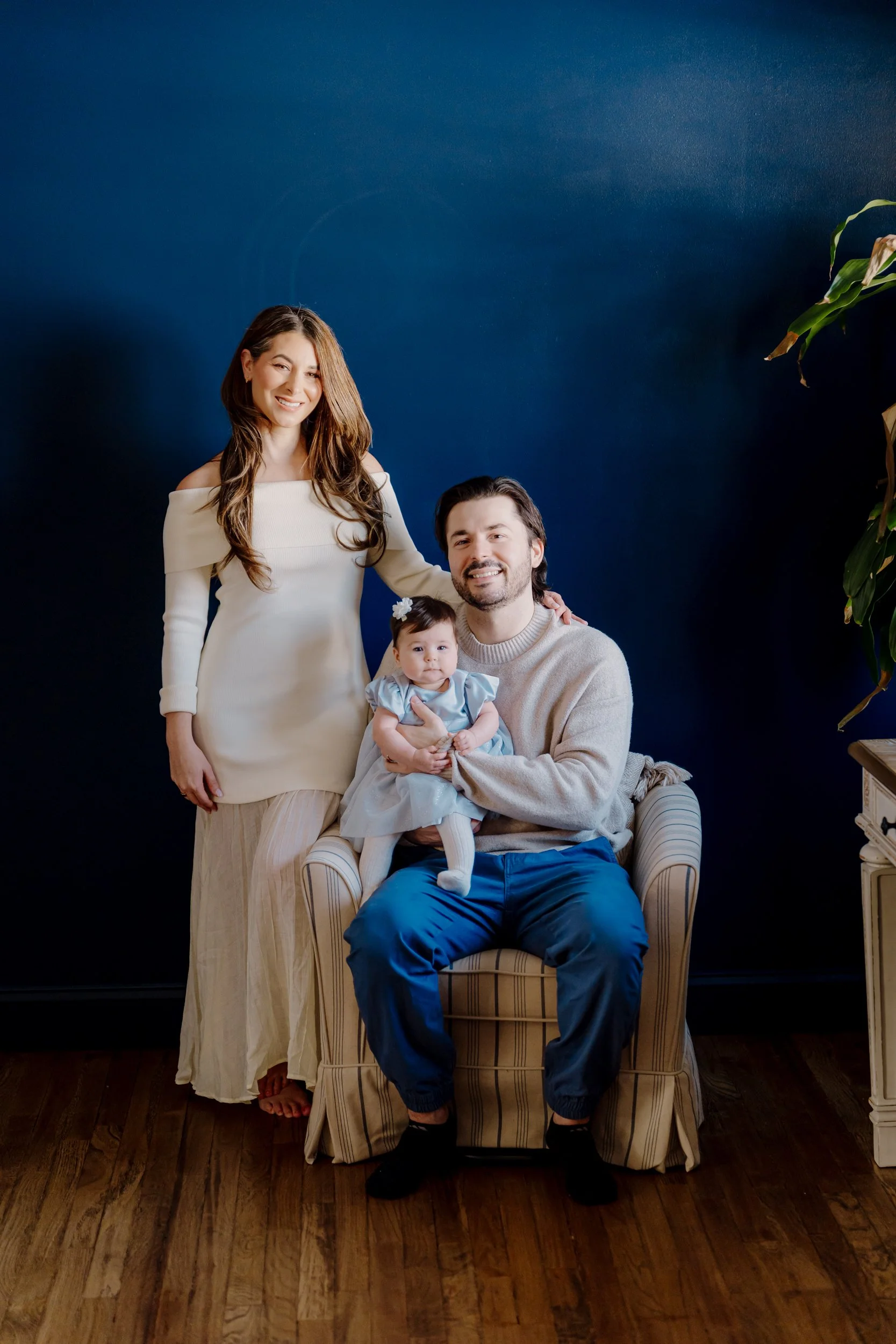 A family portrait featuring a woman in an off-shoulder cream dress, a man in a beige sweater holding a baby girl in a blue dress, all smiling against a dark blue background with a potted plant on the right.