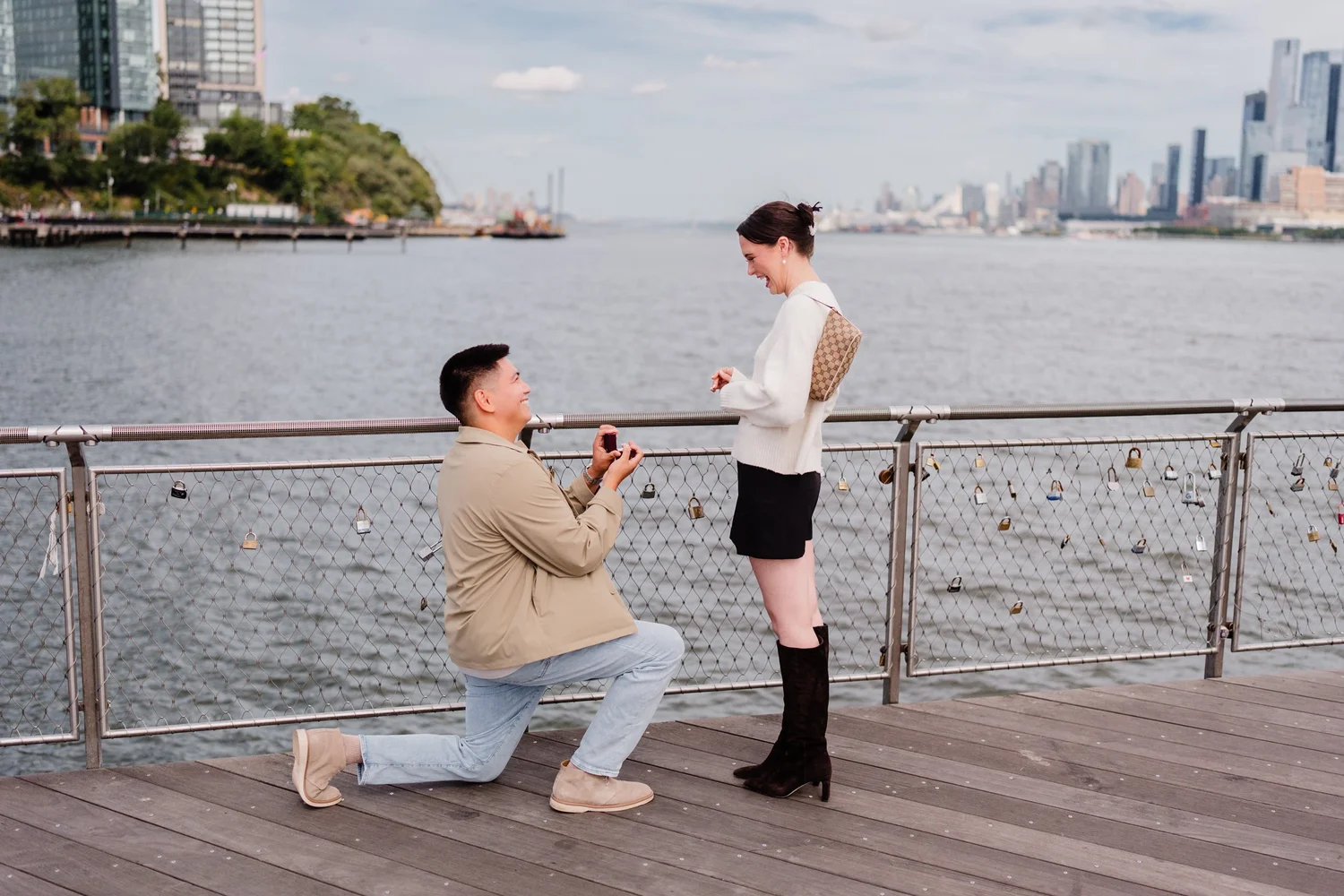A man proposes on a waterfront boardwalk with a city skyline across the water.