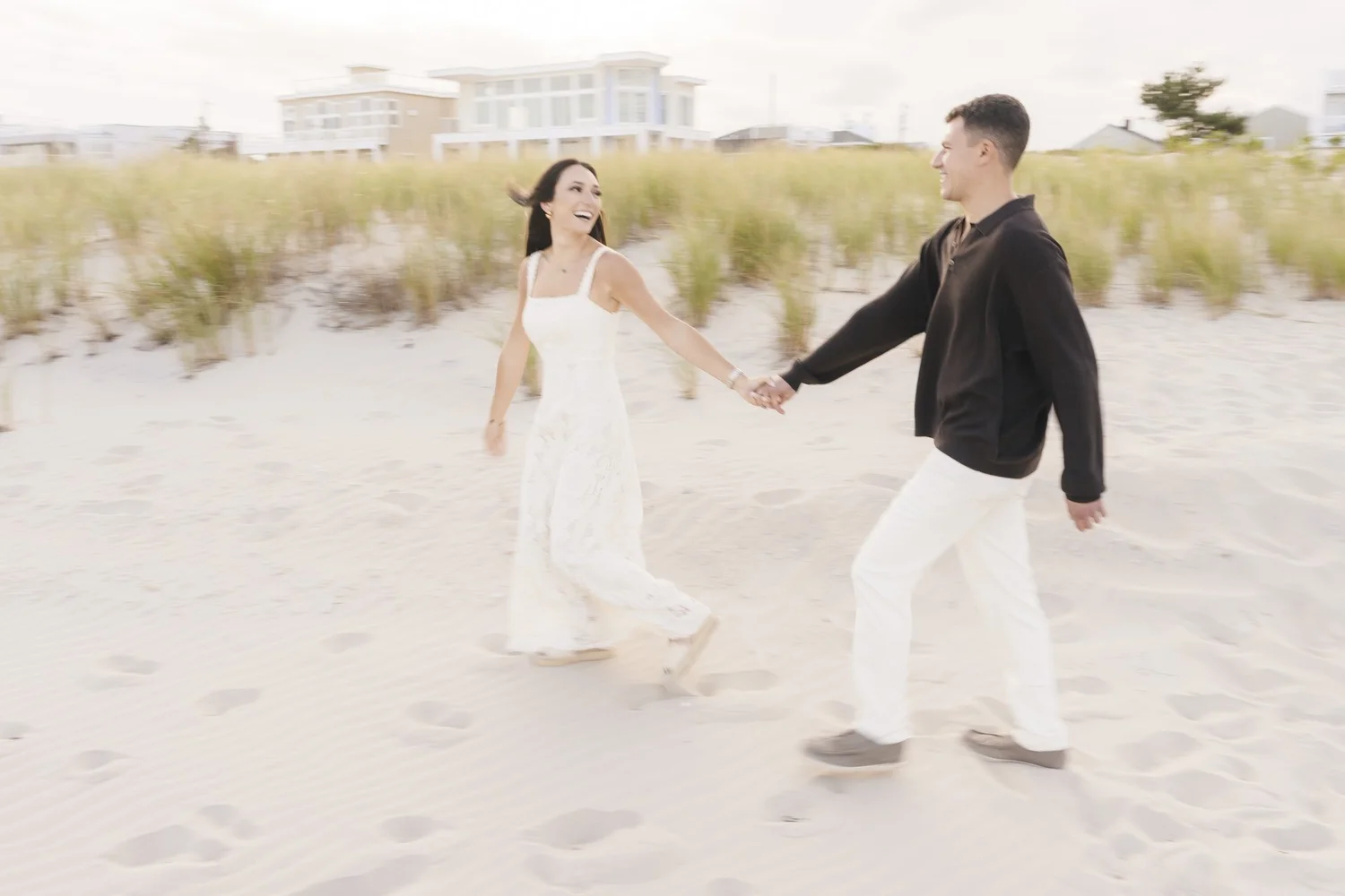 Couple holding hands and smiling while walking across the sand beside beach dunes