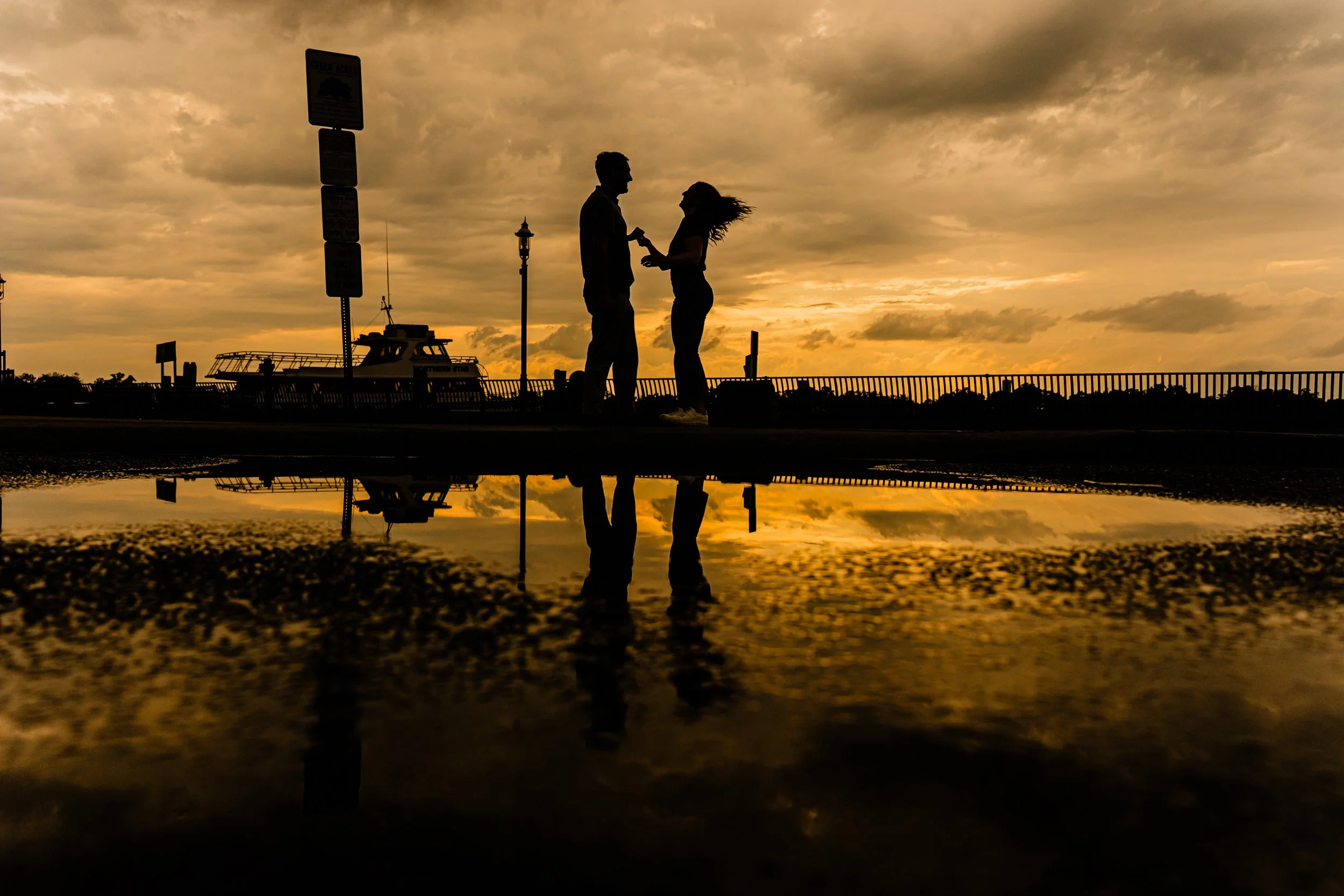 Silhouetted couple talking by the water at sunset, with clouds and a boat in the background, and their reflection visible on the wet ground.