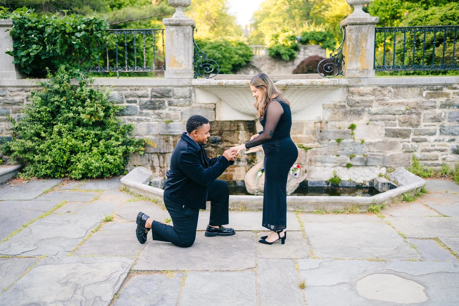A man kneels to propose beside a stone fountain as his partner smiles.