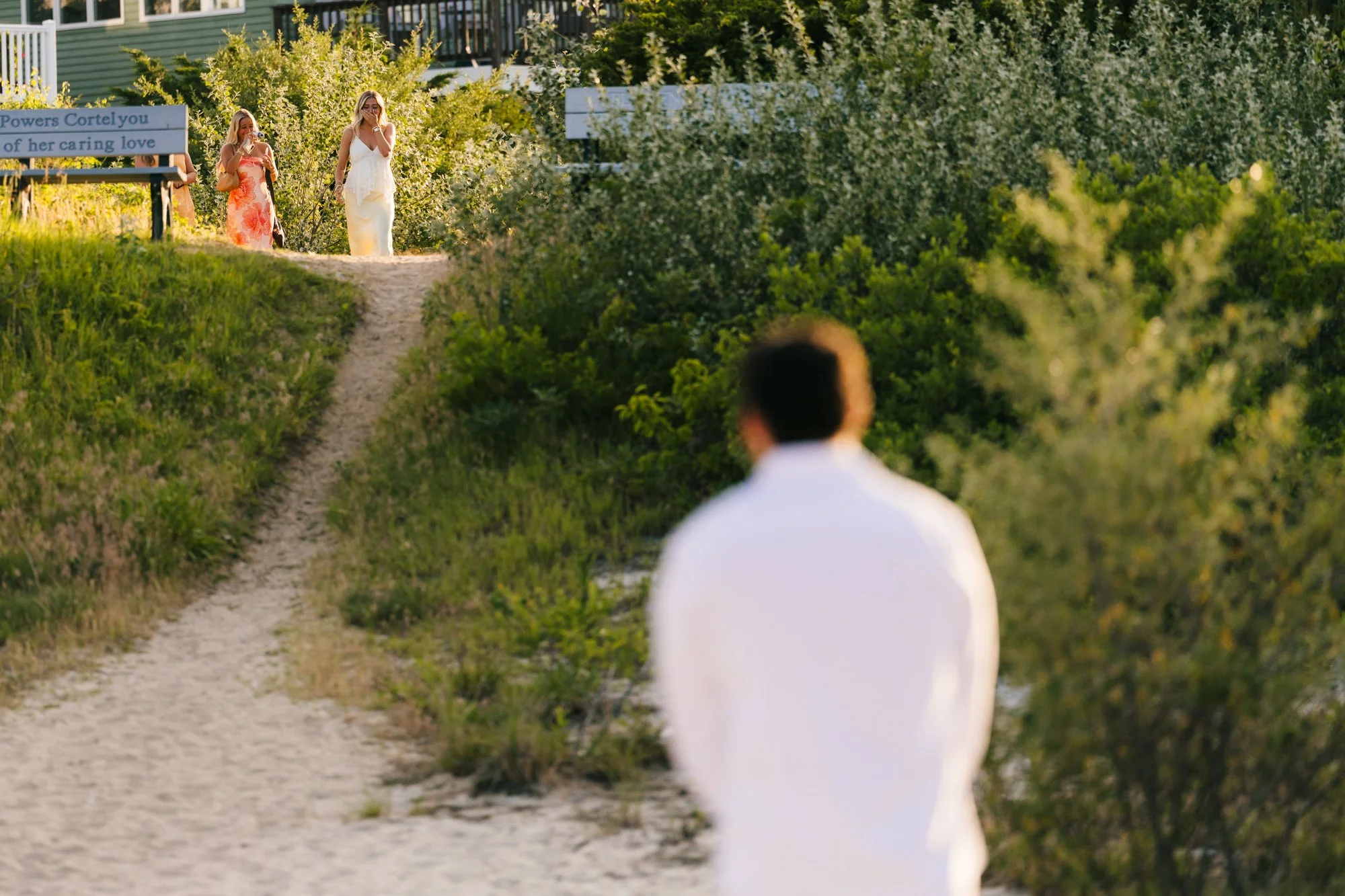 A person sitting on a bench in front of greenery, observing two women walking up a sandy path. One woman is wearing a white dress, and the other is wearing a floral dress. The scene appears to be in a garden or park area.