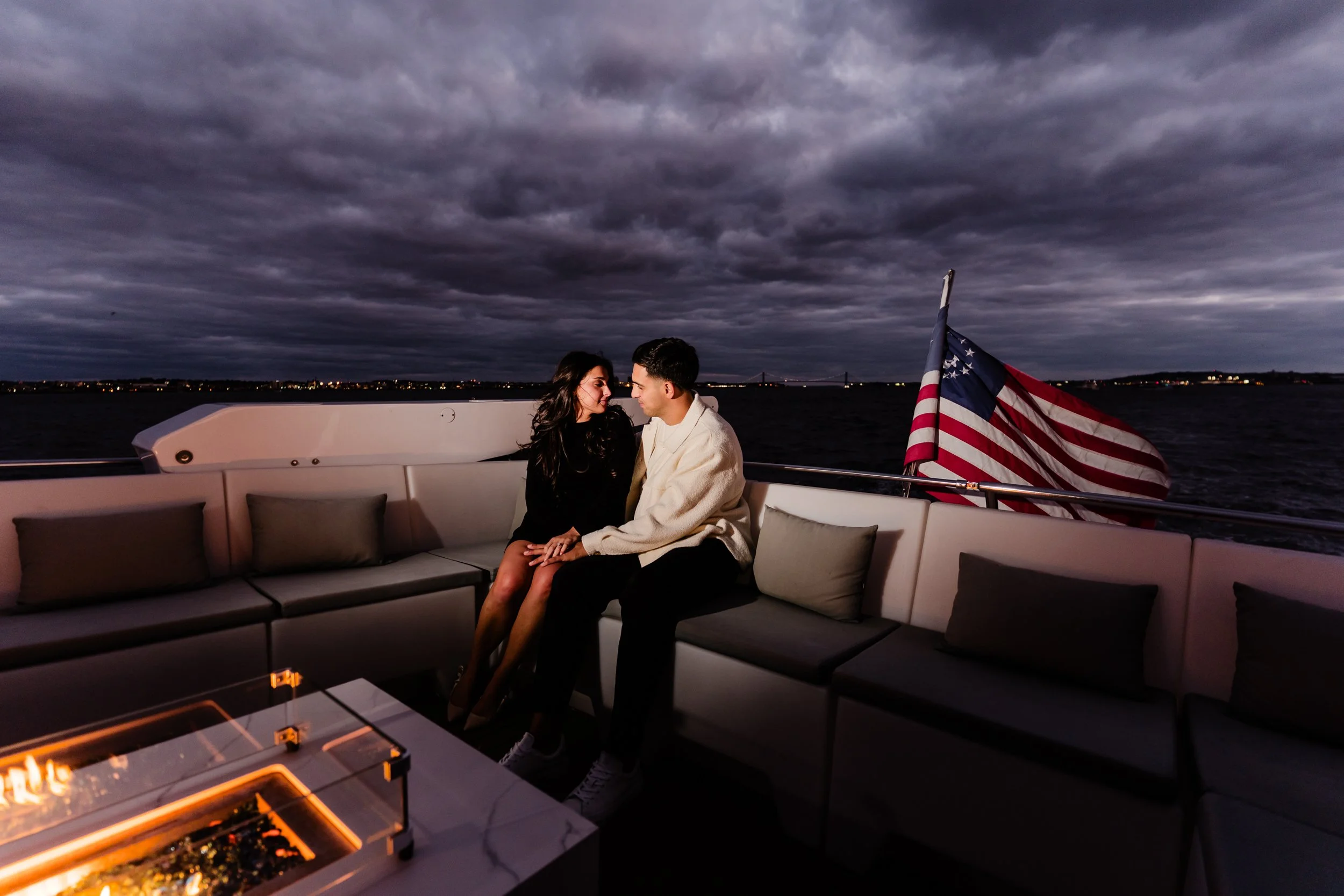 A young couple sitting closely together on a white boat at night, with a dark cloudy sky and city lights in the distance. An American flag is visible on the right side of the image.