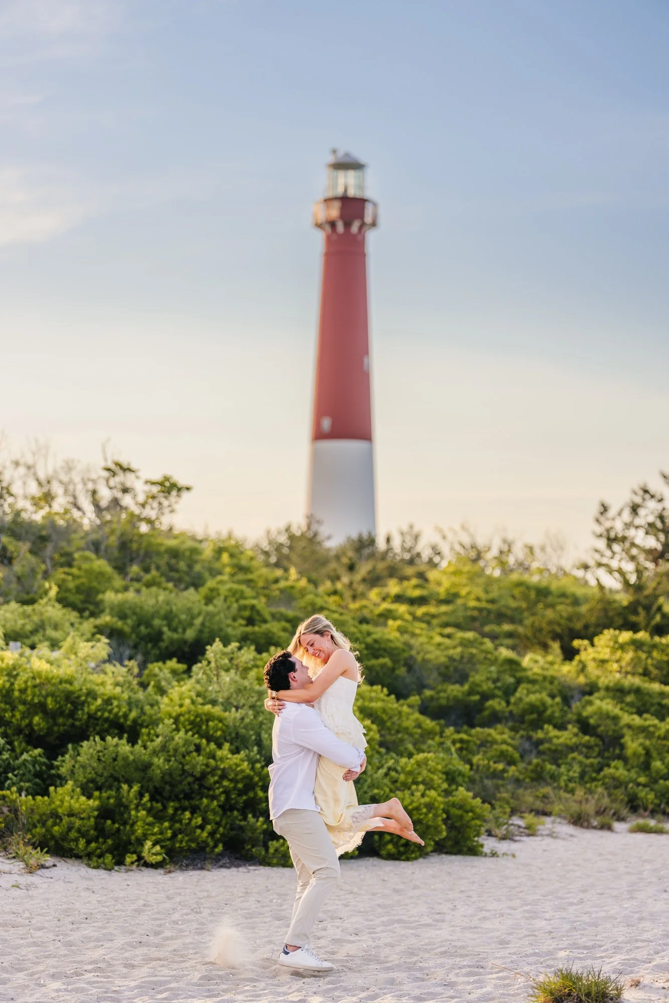A couple on a sandy beach with greenery and a red lighthouse in the background. The man is lifting the woman, who is smiling and wearing a white dress. The scene is during sunset or late afternoon.