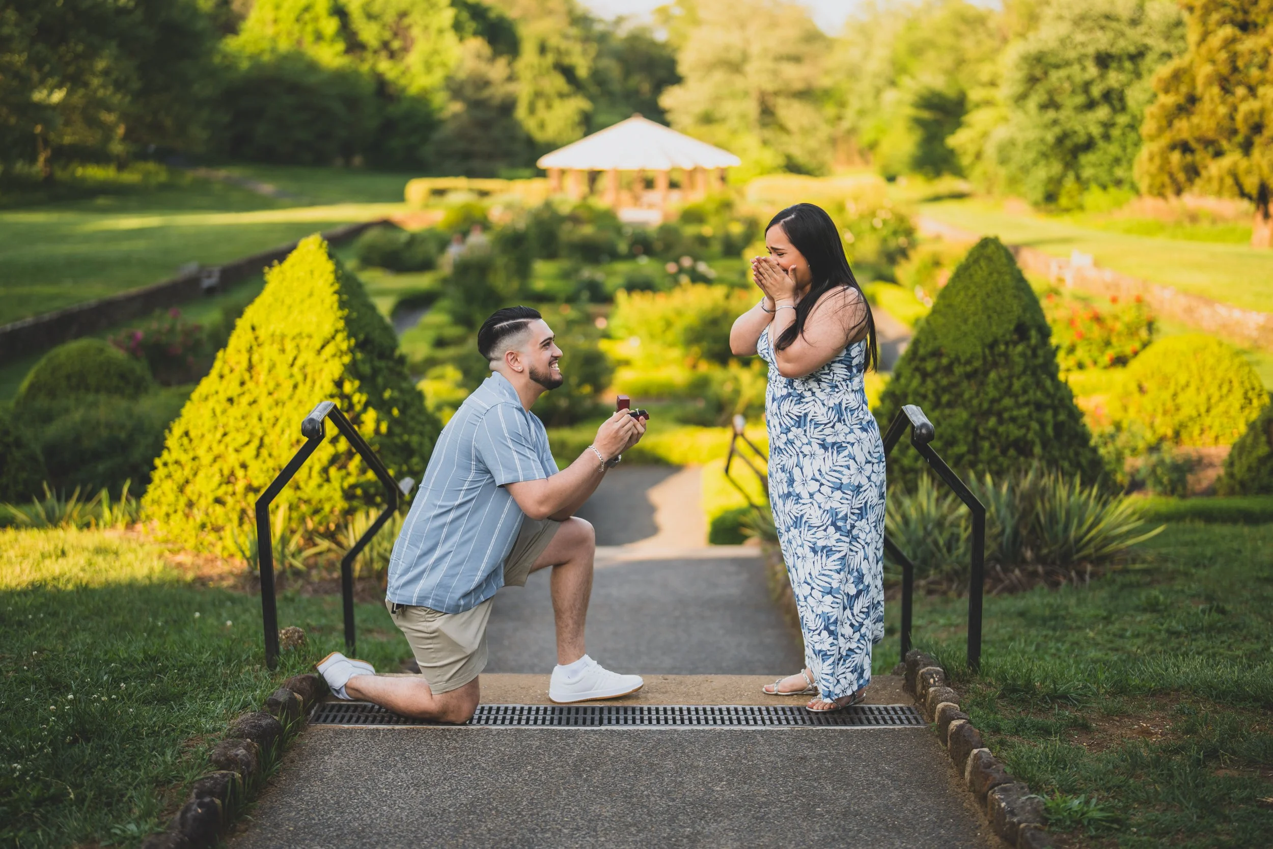 A man proposing to a woman during a marriage proposal in a garden with manicured bushes, trees, and a gazebo in the background.
