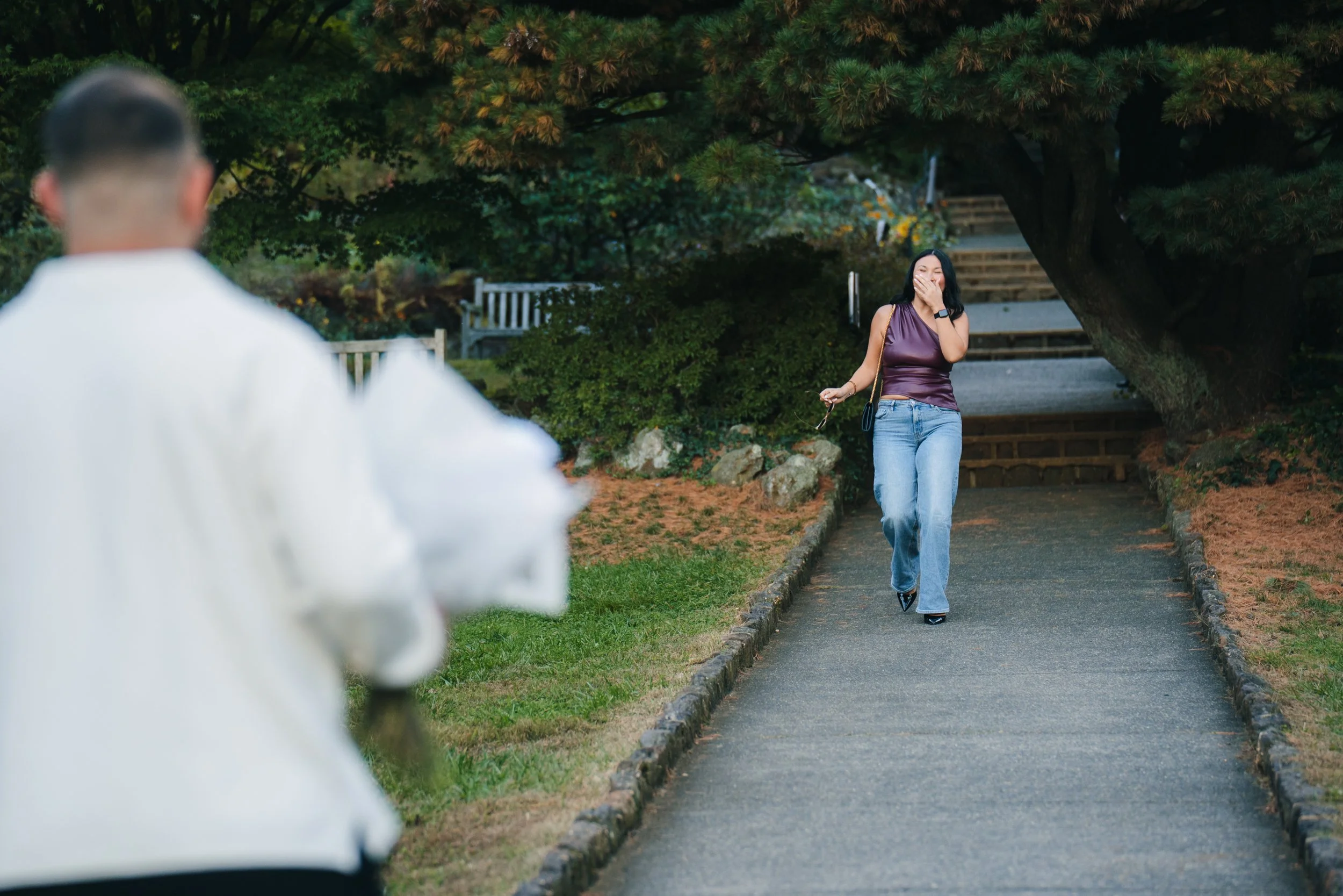 A woman in a purple top and blue jeans walking along a park path, covering her mouth with her hand and smiling, as a man in a white shirt approaches from behind.