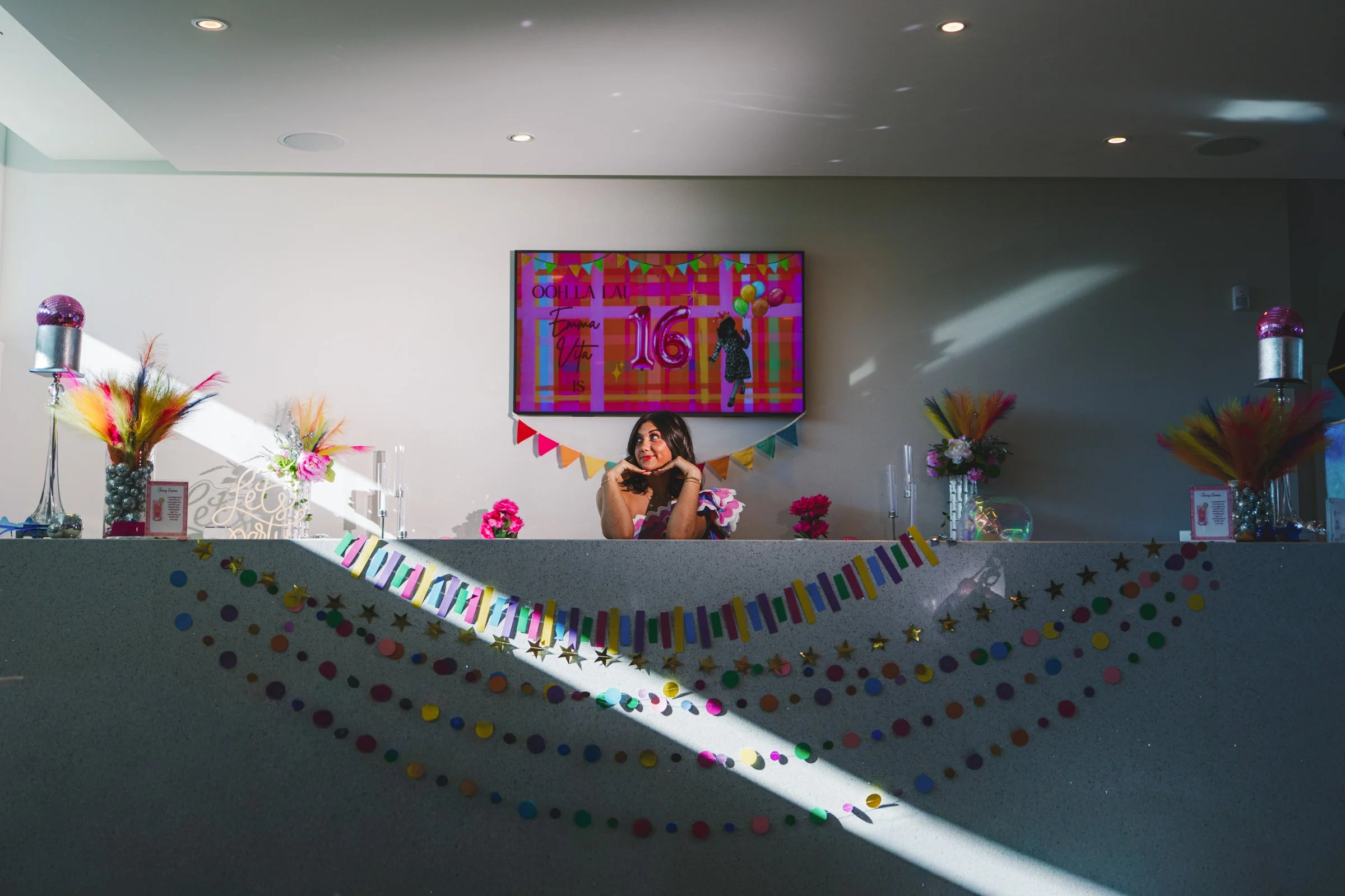 A young woman sits at a decorated party table with colorful garlands, pink flowers, and decorative vases, in a brightly lit room. A large digital screen behind her displays birthday graphics with the number 16, indicating a sweet sixteen celebration.