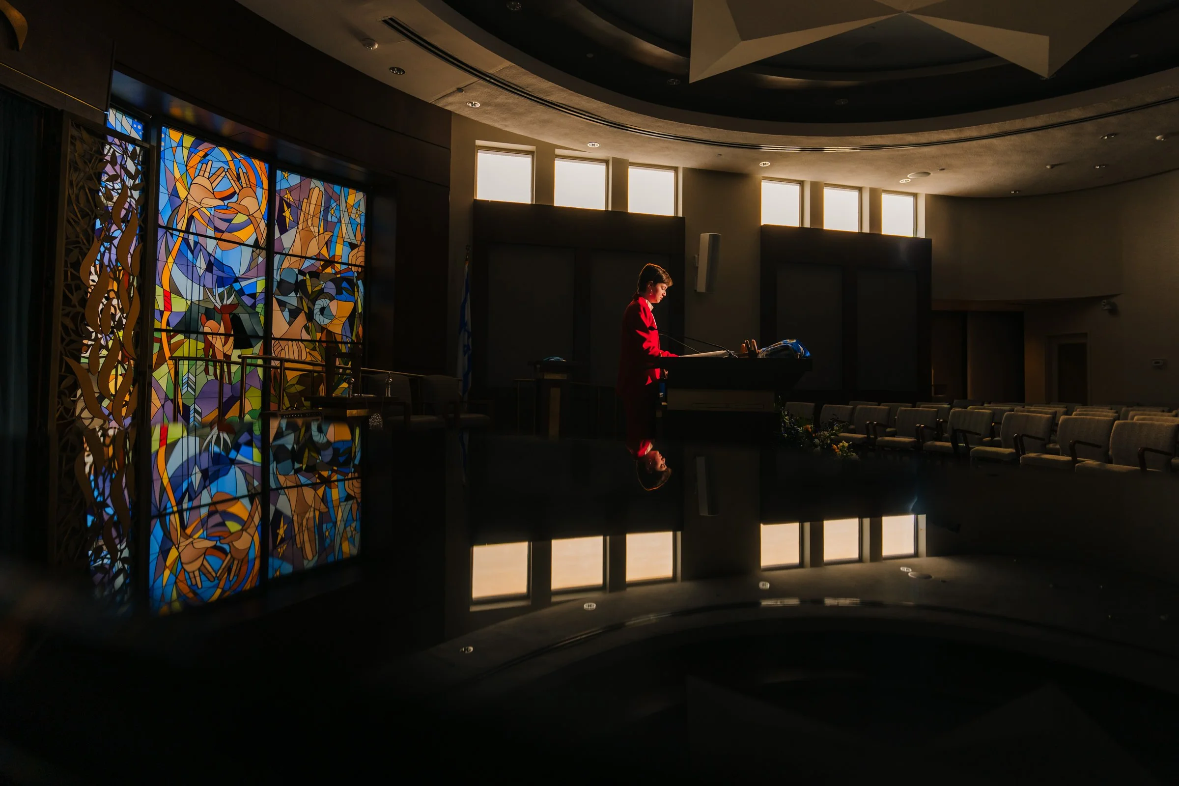 A woman in a red suit speaking at a podium in a dimly lit auditorium with large stained glass windows, empty chairs, and a reflective surface in the foreground.