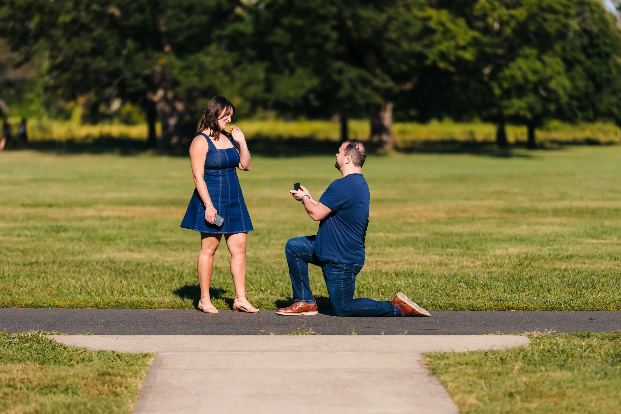A man on one knee proposes to a woman in a park.
