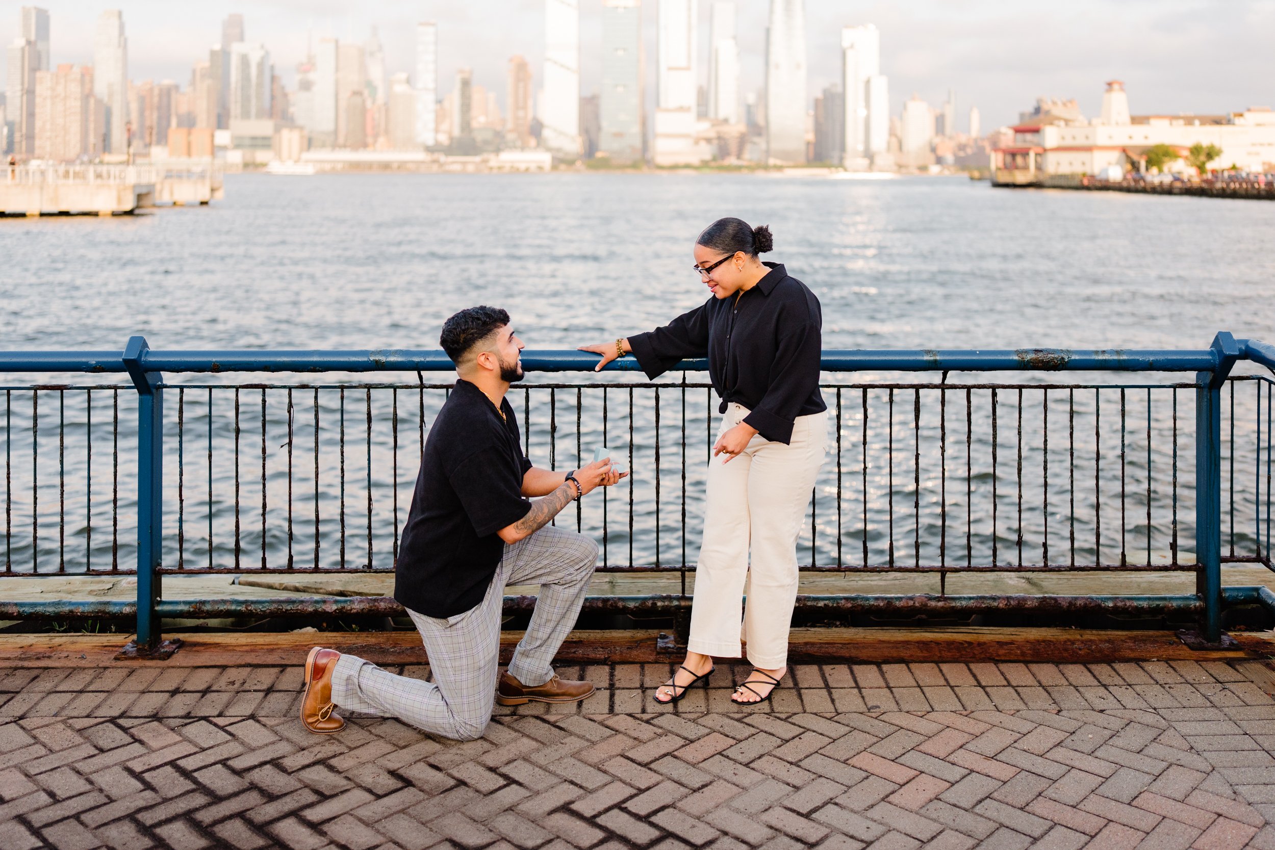 A man is kneeling on one knee proposing to a woman by a body of water with a city skyline in the background. The woman is standing, smiling, and looking down at the man. They are near a metal railing on a paved walkway.
