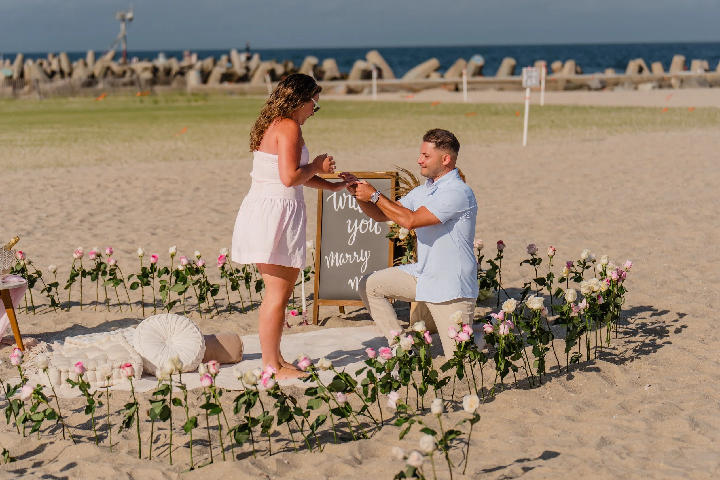 A man proposes to a woman during a beach wedding ceremony with flowers, cushions, and a sign that says "Will You Marry Me?" in the background, near the ocean and a breakwater.