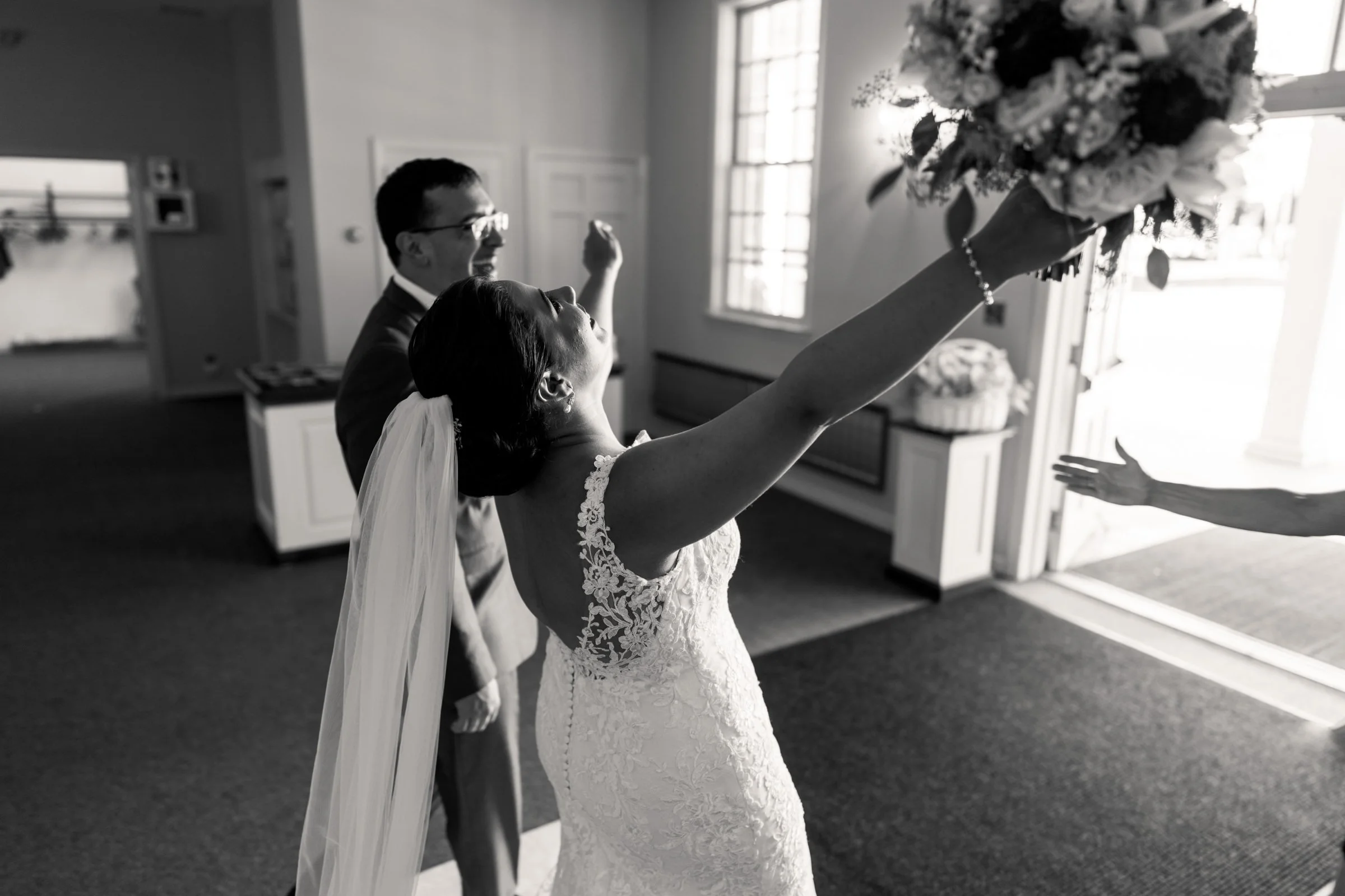 Black and white photo of a bride in a wedding dress and veil raising a bouquet, with a smiling groom in a suit behind her, inside a bright room.