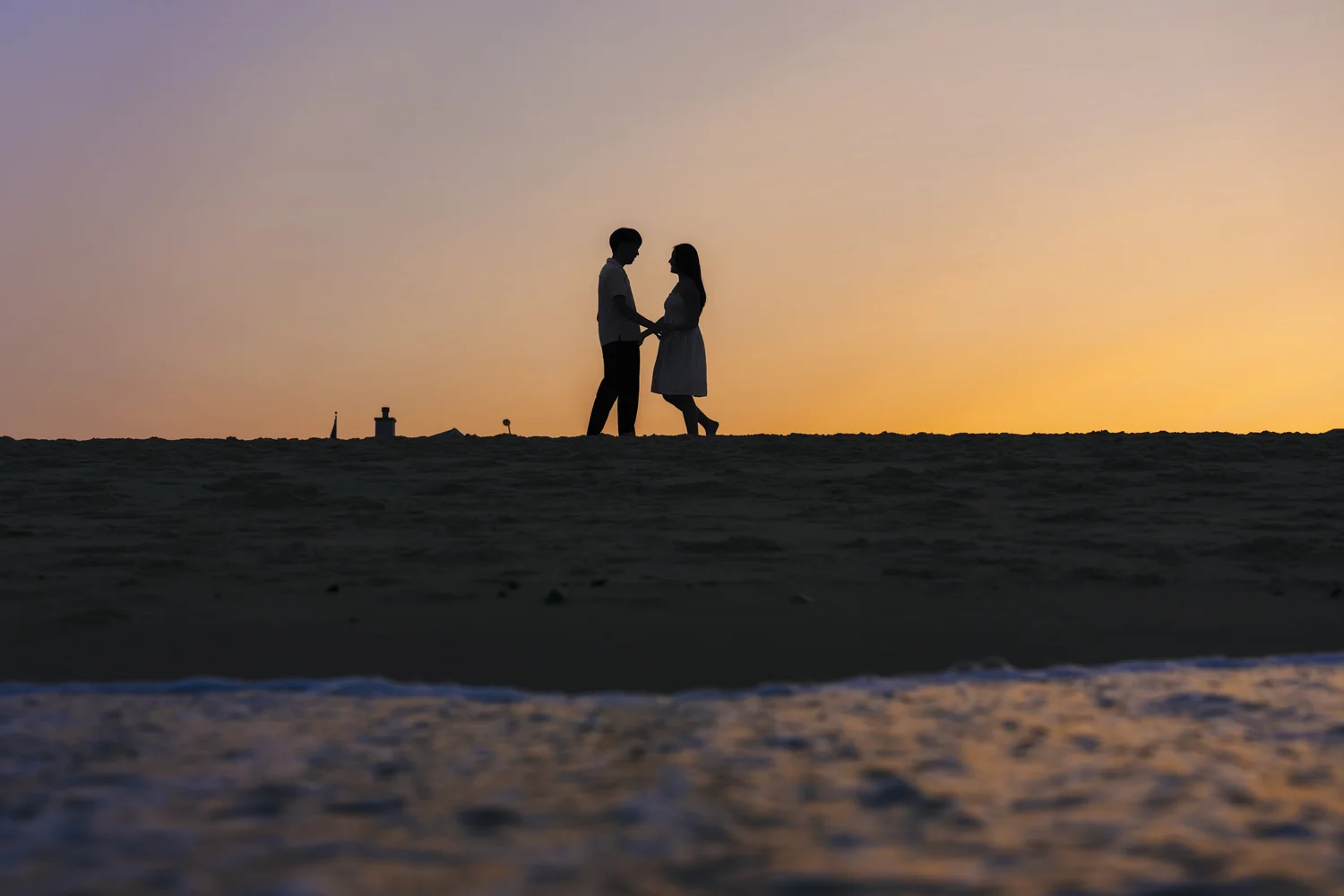 Silhouettes of a couple facing one another on the beach against an orange sunset.