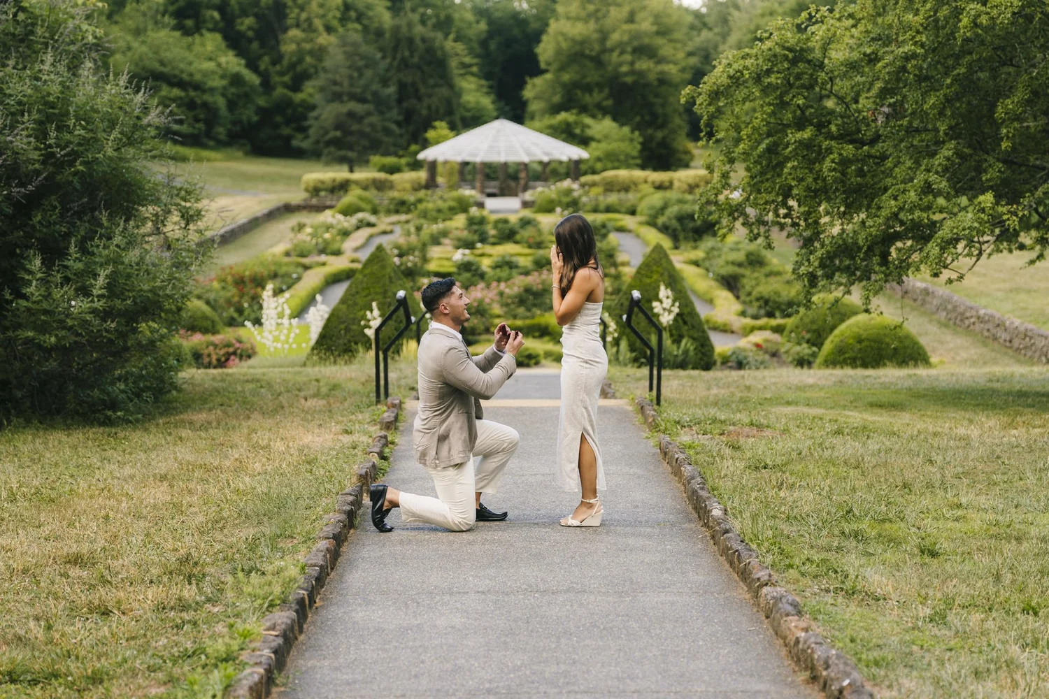 Man proposing on a garden path overlooking formal flower beds and a gazebo