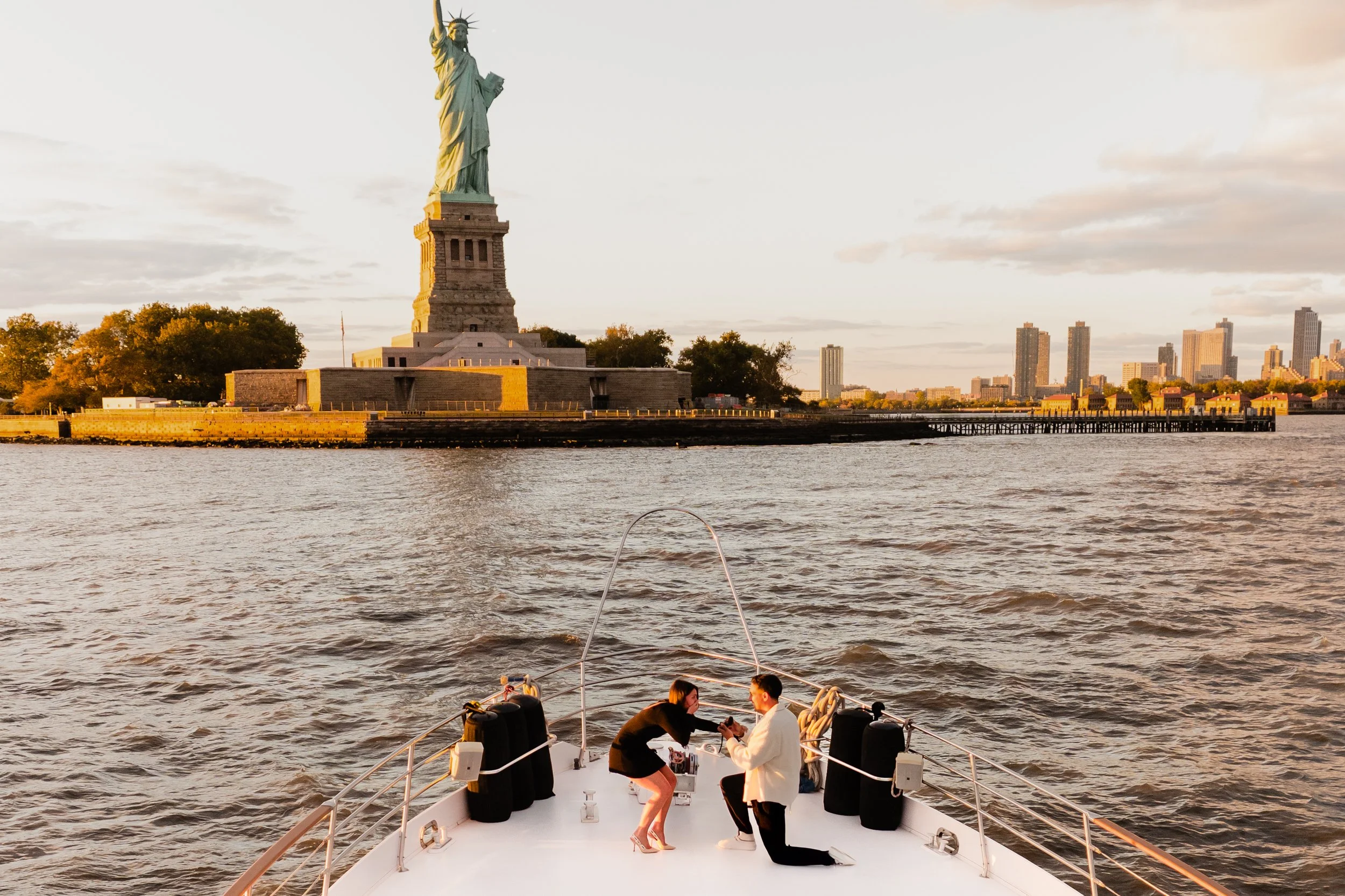 People on a boat, with the Statue of Liberty and New York City skyline in the background during sunset.
