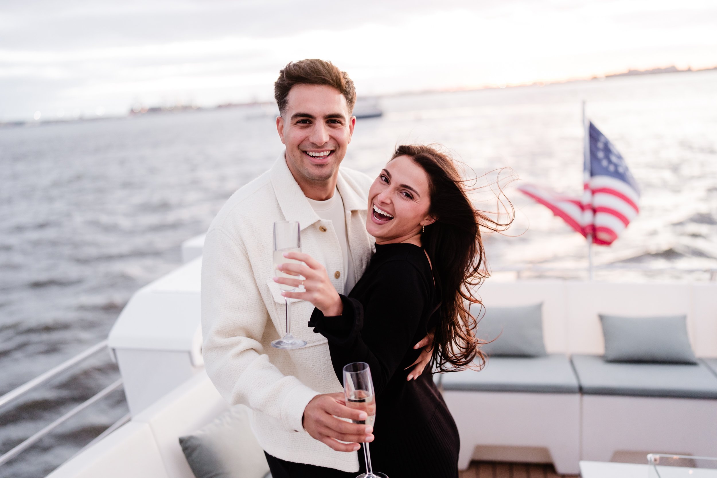 A happy young couple on a boat, smiling at the camera, holding glasses of champagne, with an American flag in the background on the right, and water and cloudy sky in the distance.