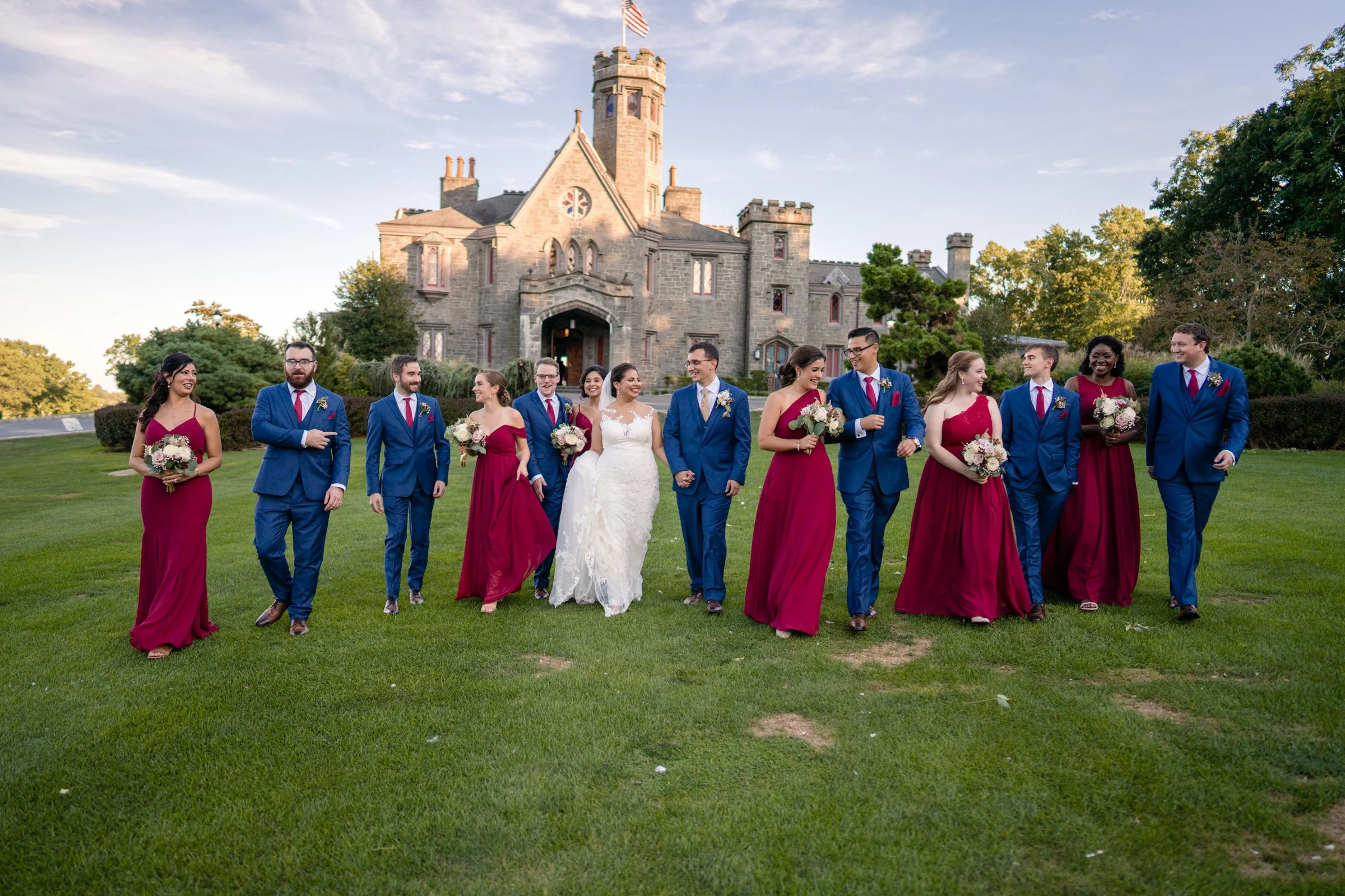 A wedding party walking on a grassy lawn outside a castle-like building during daytime. The group includes a bride in a white gown, bridesmaids in maroon dresses, and groomsmen in blue suits.